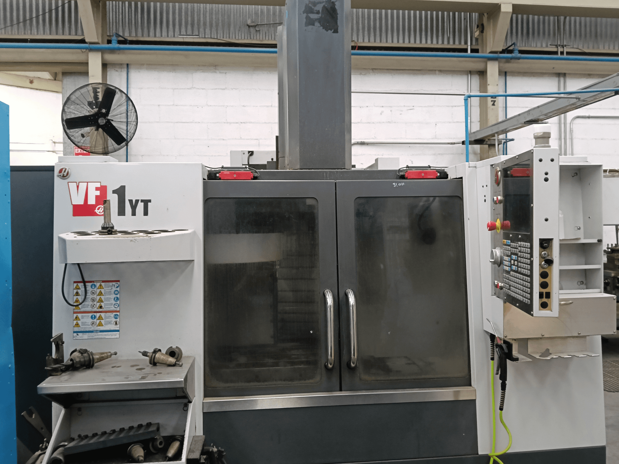 A HAAS VF-1YT-1 függőleges megmunkáló központ elölnézete, oldalán a vezérlőpanellel és a szerszámokkal.