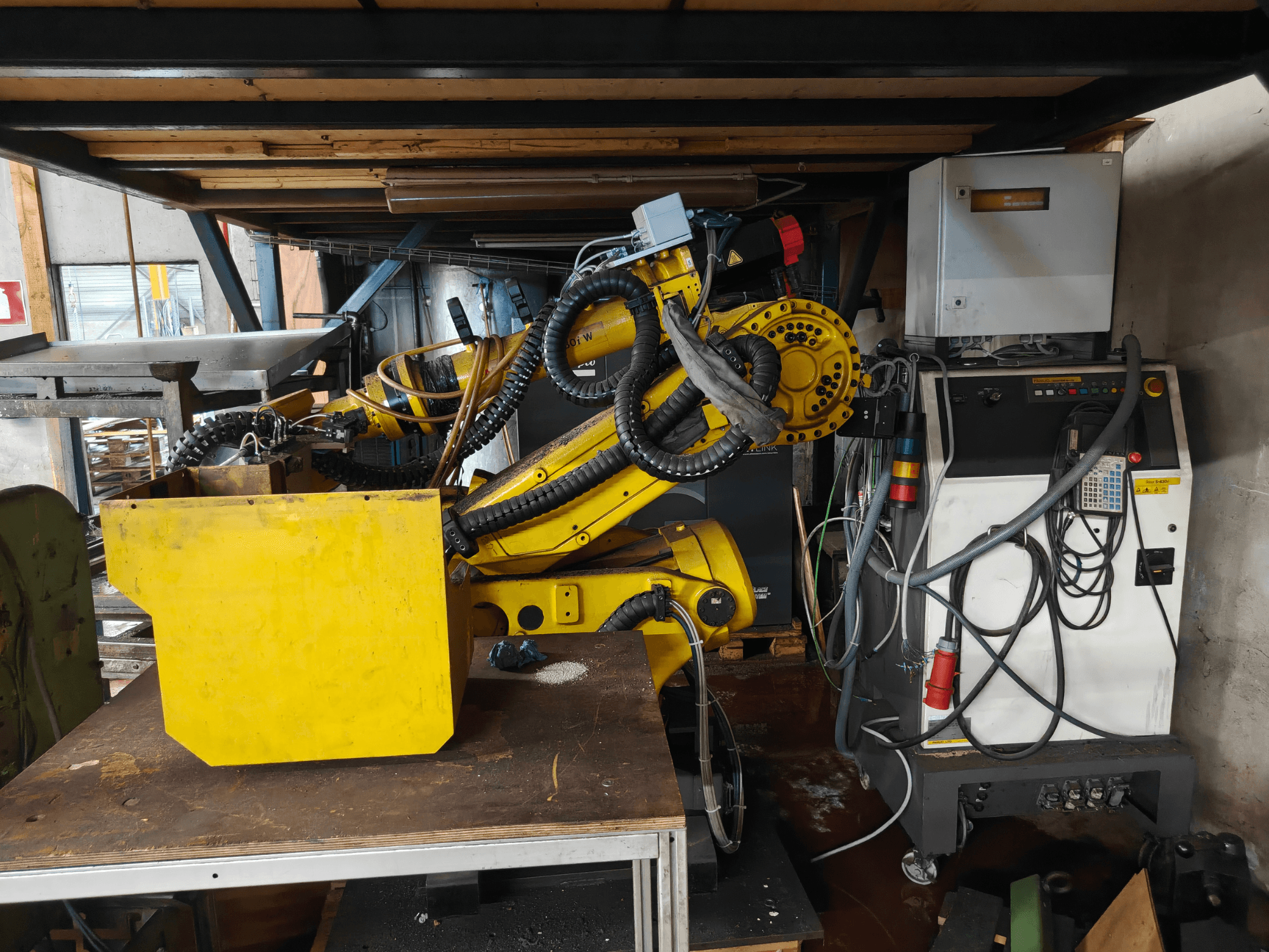 A FANUC S-430i W robotkar elölnézete: sárga színű, vezérlőpanellel, kábelekkel és egy sárga alkatrésszel a munkapadon.
