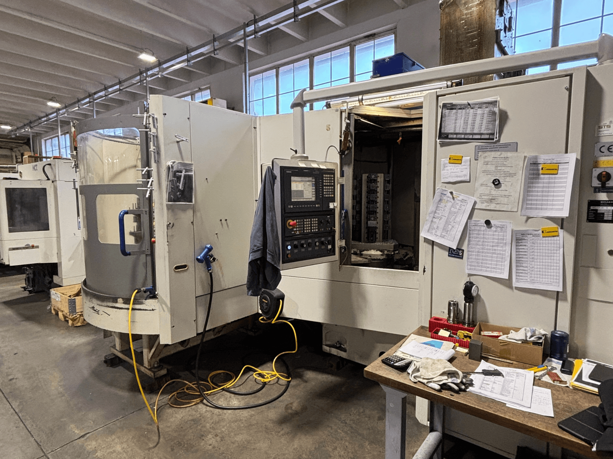 CNC-gép egy műhelyben, elölnézetben, a vezérlőpanel és a dokumentáció az oldalon; szerszámok és anyagok az asztalon.