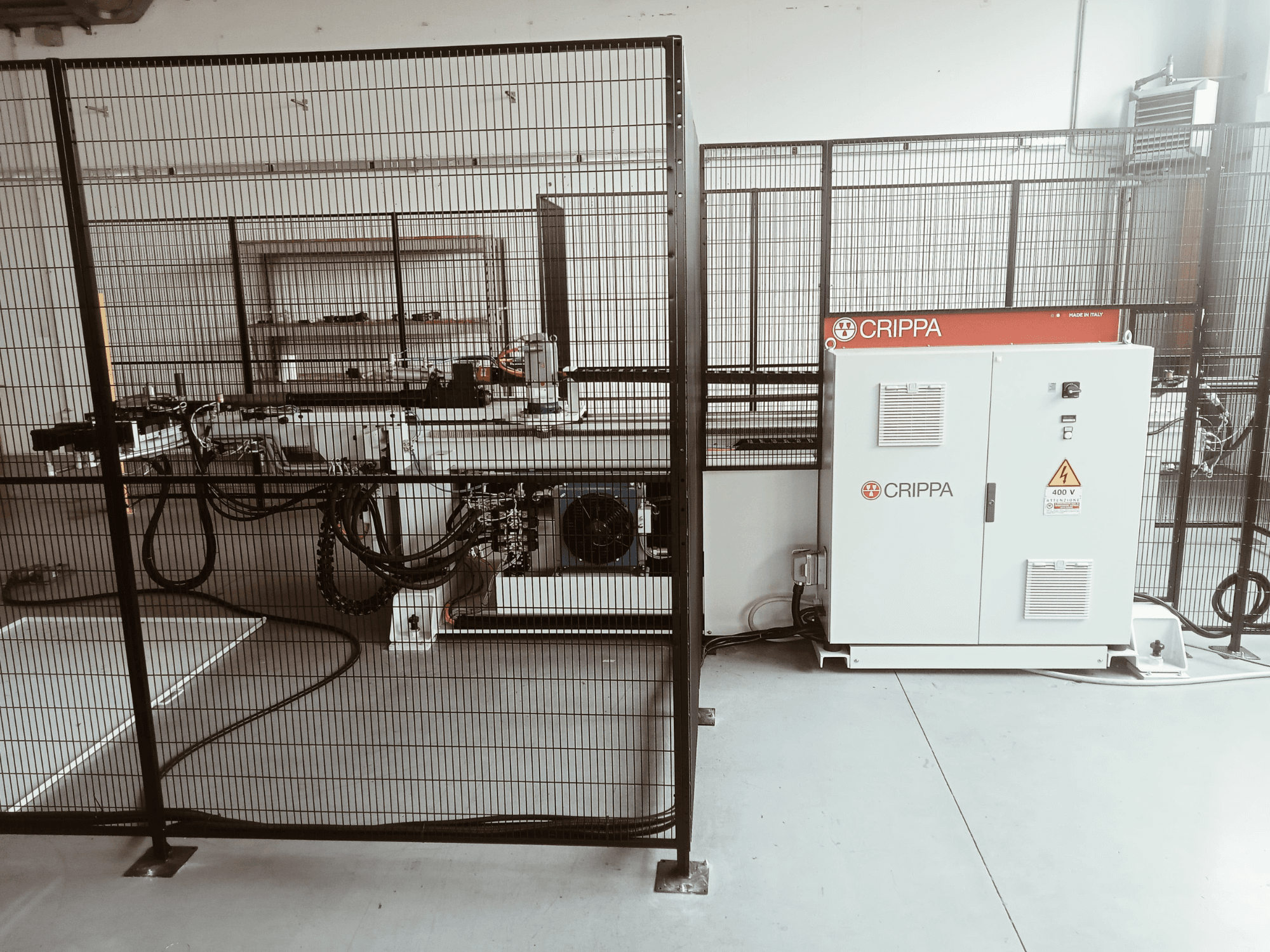 Fehér CRIPPA gép egy biztonsági ketrec mögött, látható tömlőkkel és alkatrészekkel, oldalnézet, ipari környezetben.