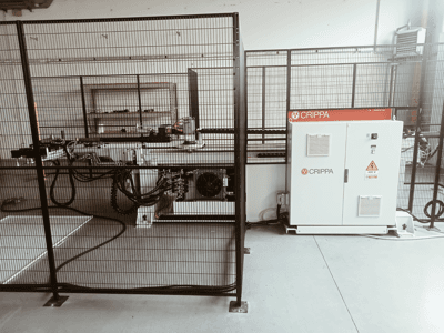 Fehér CRIPPA gép egy biztonsági ketrec mögött, látható tömlőkkel és alkatrészekkel, oldalnézet, ipari környezetben.