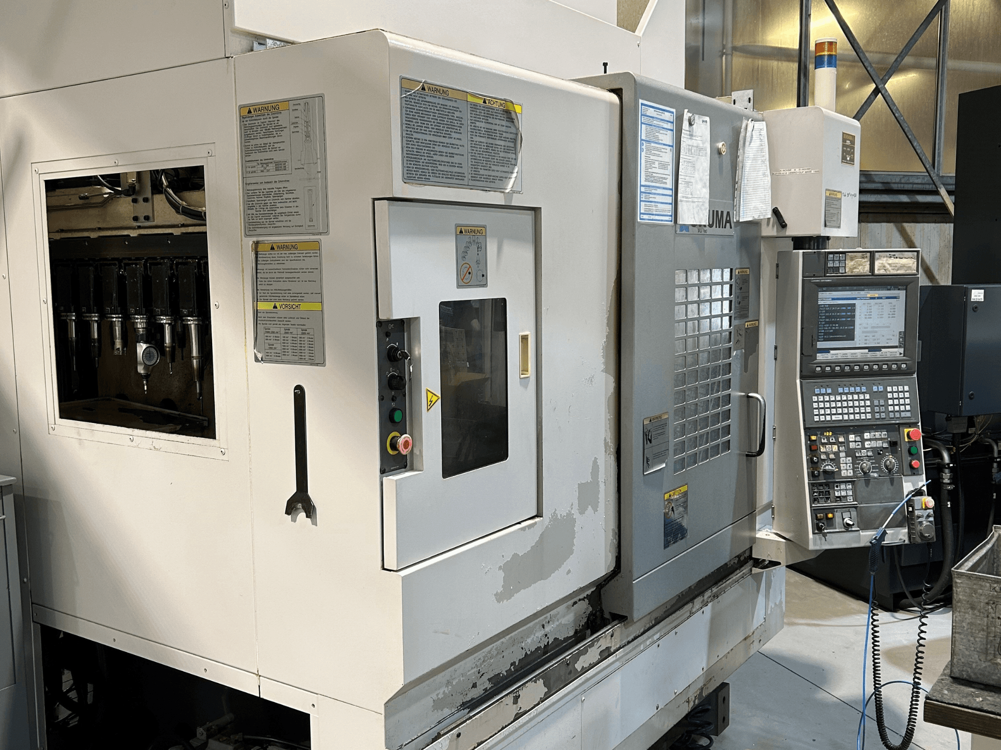 Egy UMA CNC-gép előlapja látható szerszámtartókkal, vezérlőpanellel, figyelmeztető feliratokkal és kezelőgombokkal.