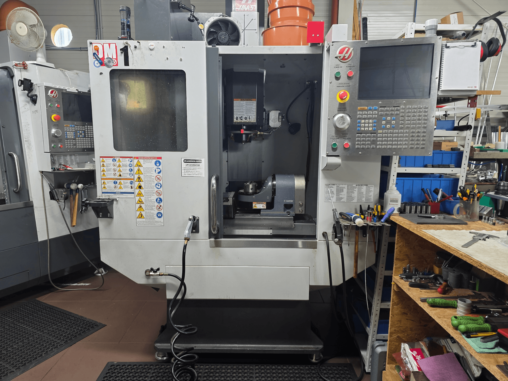 Haas CNC-gép modell VF-2, előlnézet, a vezérlőszervek, az orsó területe, a biztonsági címkék és a szerszámok egy közeli munkapadon.