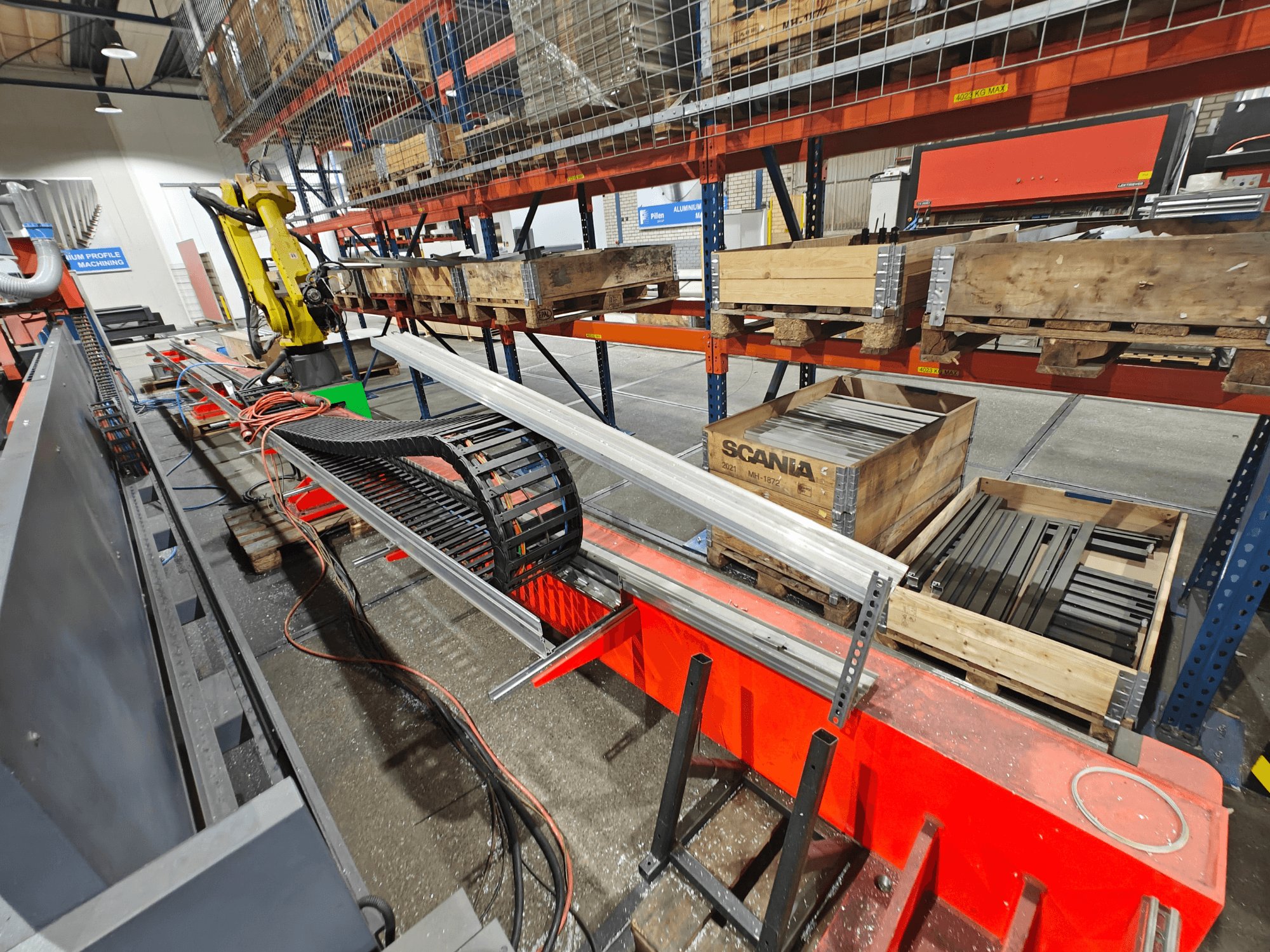 Robotkar a munkaállomáson, narancssárga és fekete kábelek láthatóak, Scania ládákkal és alumínium profilokkal egy gyárban.