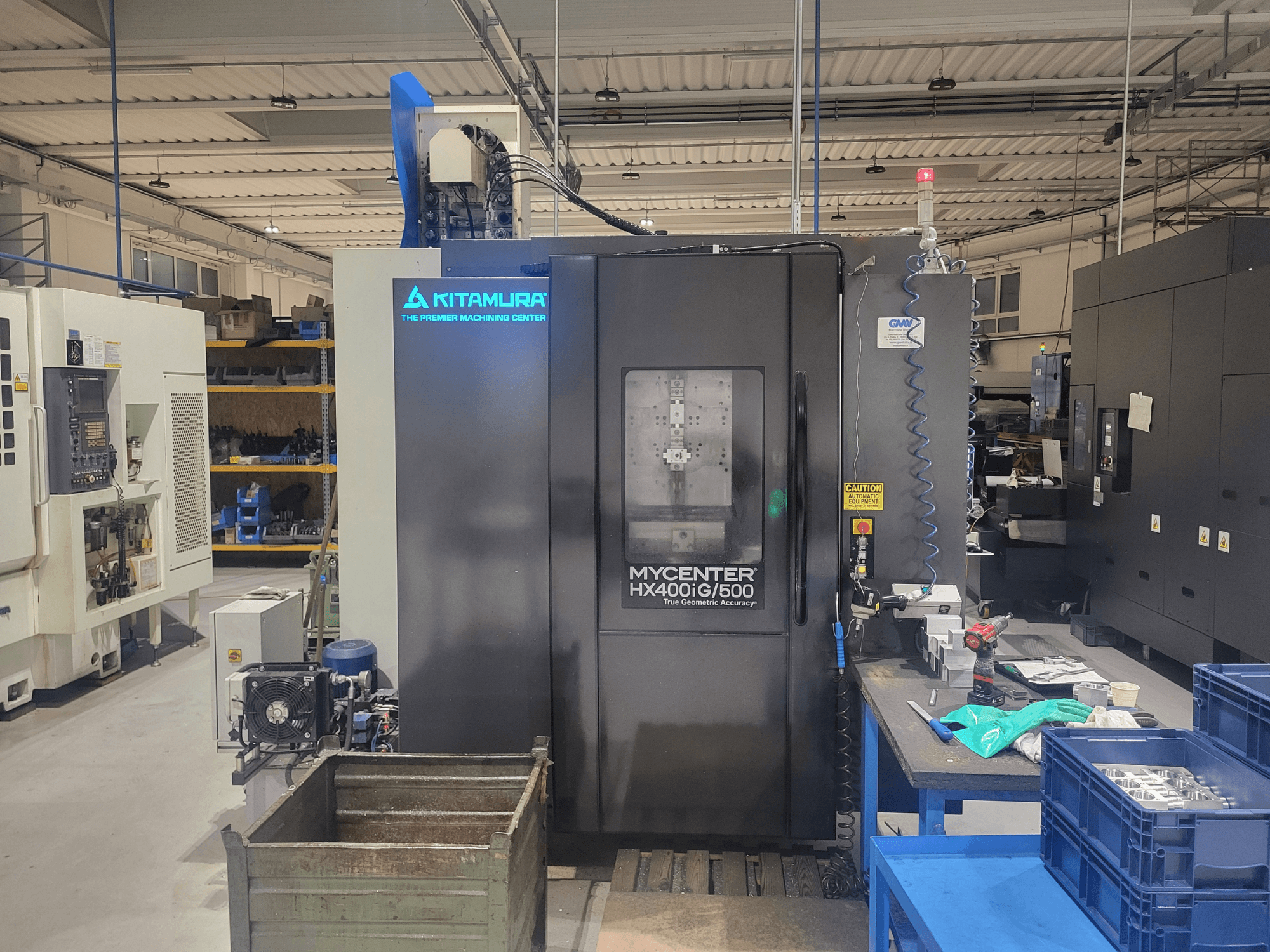 Kitamura Mycenter HX400iG/500 CNC-gép egy műhelyben, elölnézetben, a kezelőszervekkel, a szerszámtárolóval és az anyagokkal a közelben.