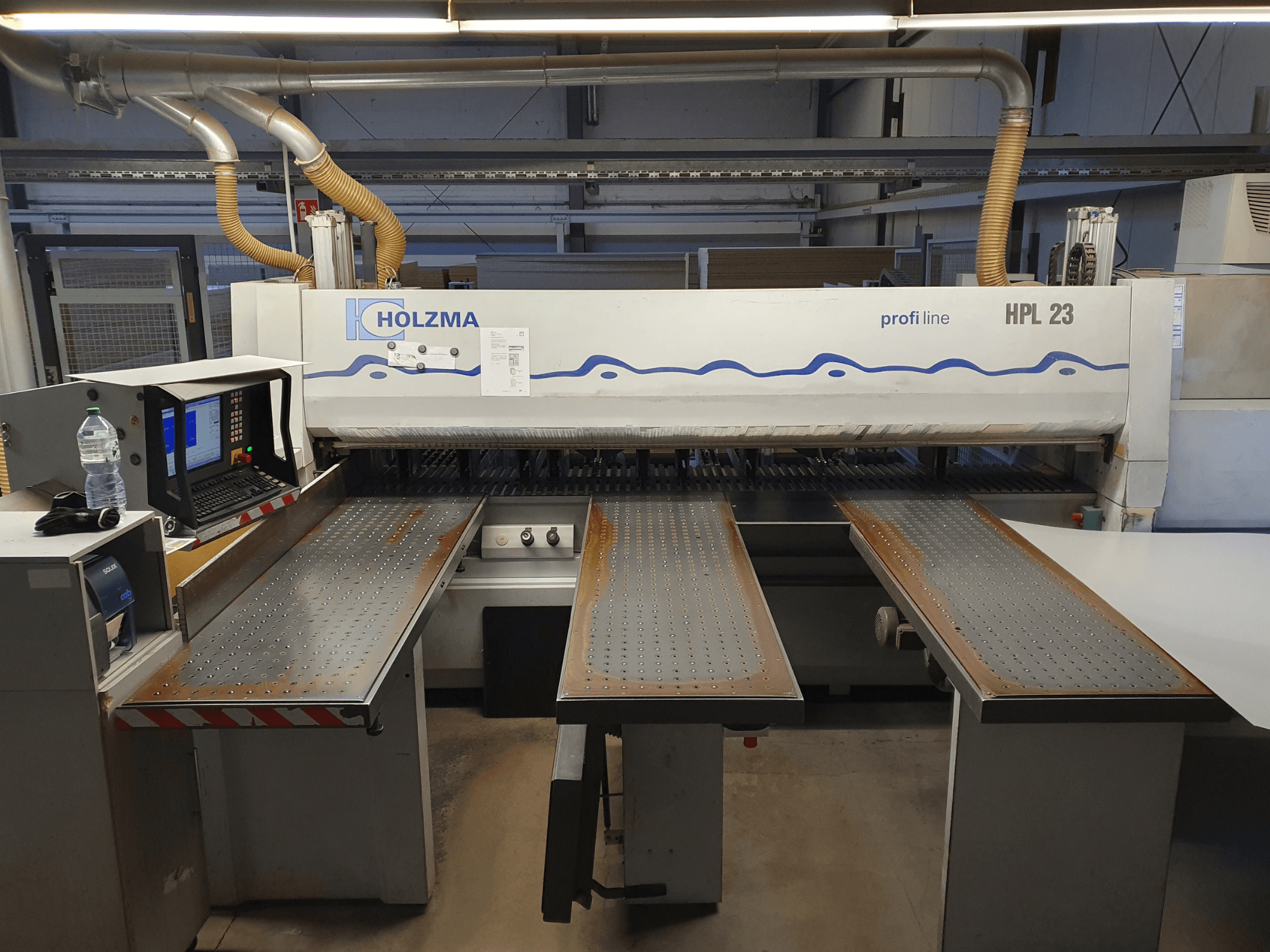 HOLZMA HPL 23 vágógép szemből, három perforált asztallal, számítógépes terminállal és vizes palackkal a közelben.