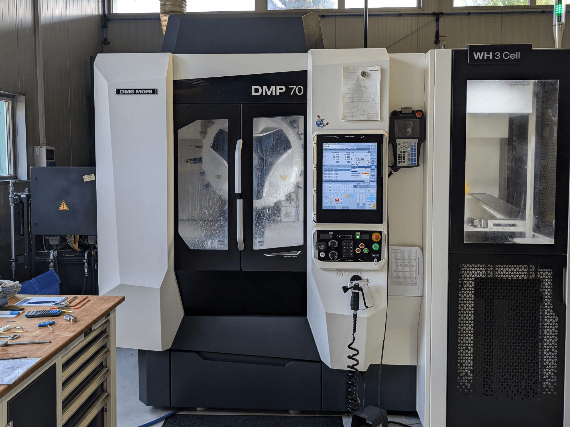 DMG MORI DMP 70 függőleges megmunkáló központ, elölnézet a vezérlőpanellel és a kezelőképernyővel, műhelyszerszámokkal körülvéve.