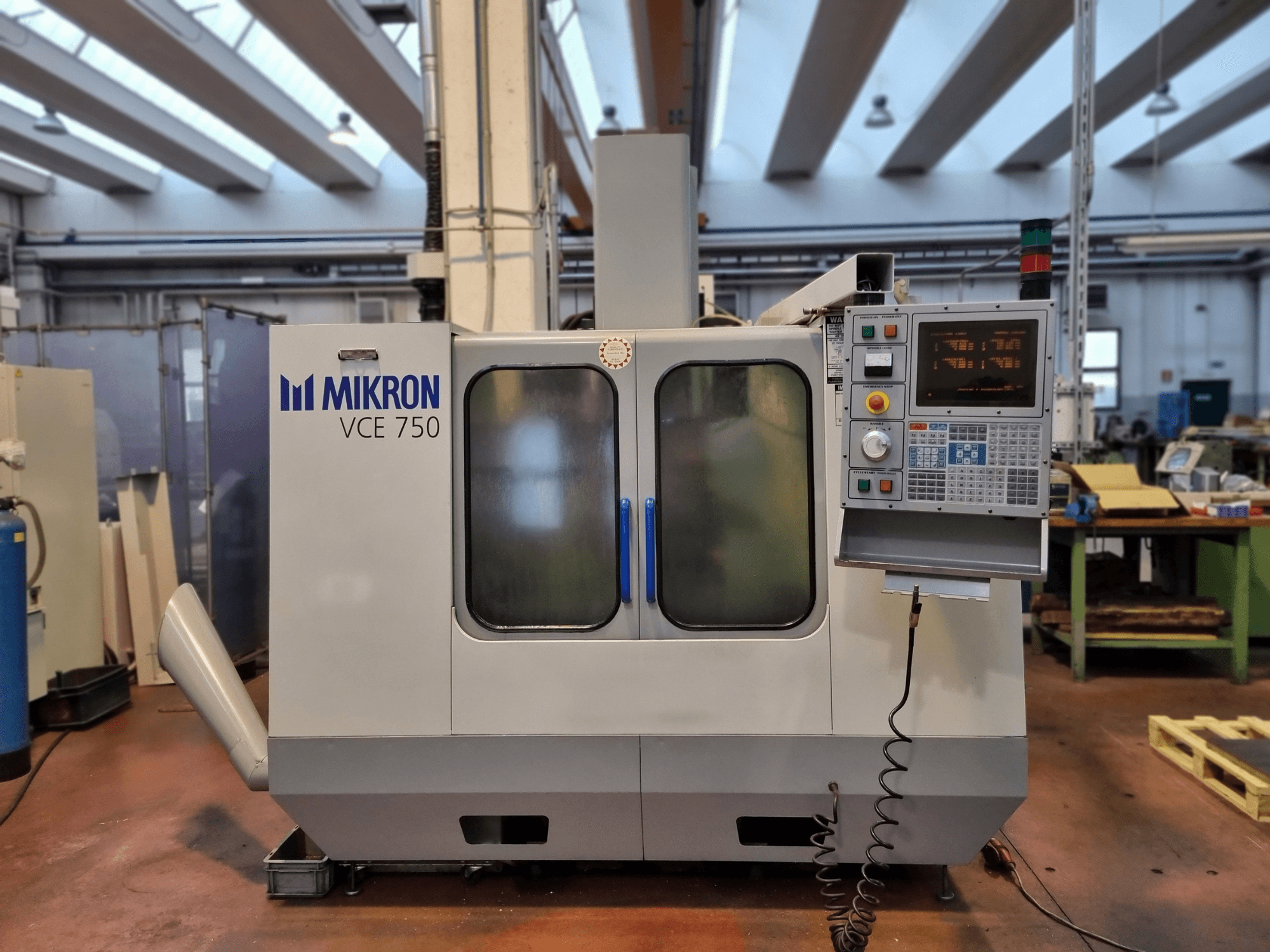 Fehér Mikron VCE 750 CNC-gép egy műhelyben, oldalnézetben, digitális vezérlőpanellel és nagy üvegajtókkal.