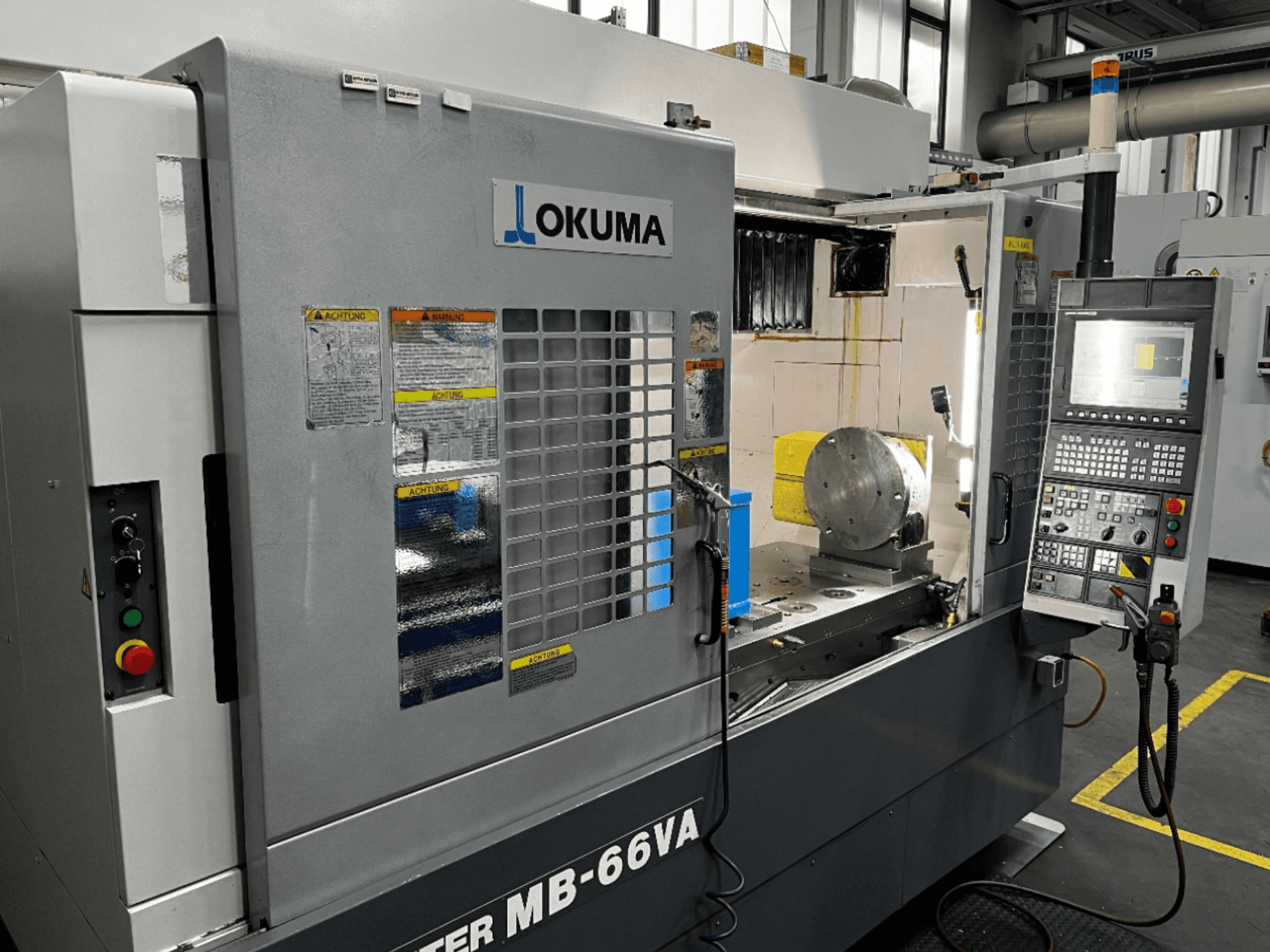 Okuma MB66-VA függőleges megmunkálóközpont előlapja, amely a vezérlőpanelt, a munkadarabot és a gép belsejét mutatja a biztonsági címkékkel.