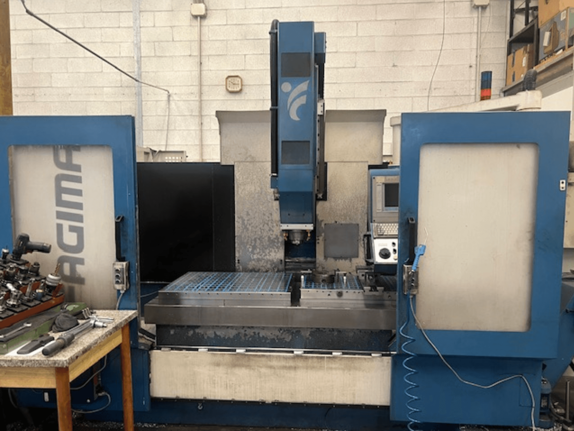 Egy JAGIMA CNC-gép elülső nézete egy nagy fém munkaterülettel, látható vezérlőpanellel és a közeli asztalon lévő szerszámokkal.