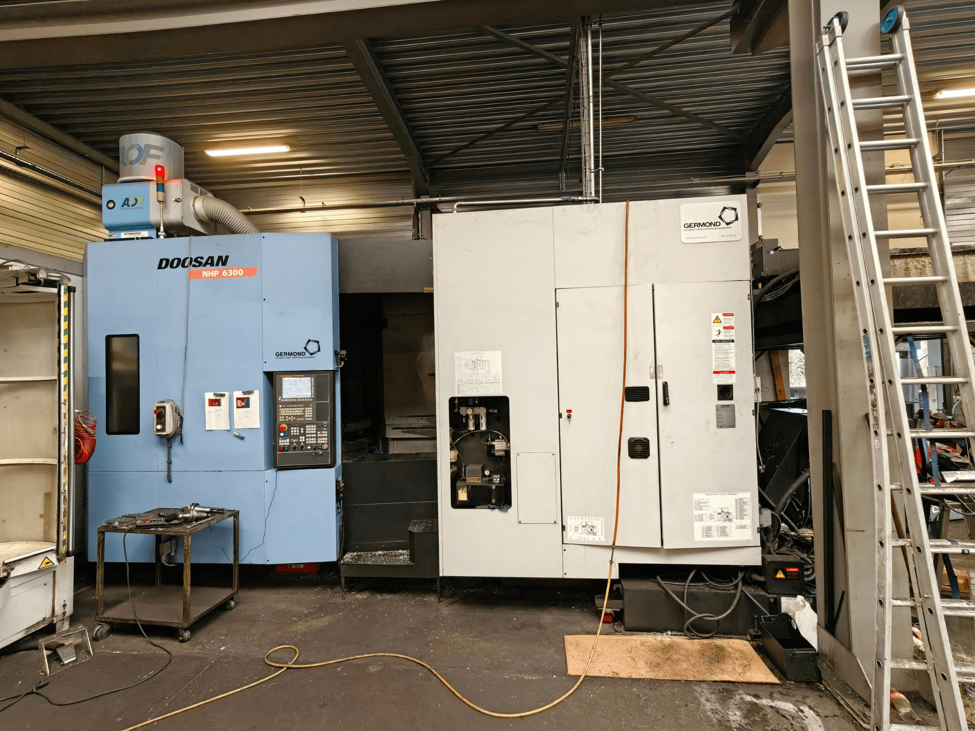 A DOOSAN NHP6300 vízszintes megmunkáló központ elölnézete, amelyen látható a vezérlőpanel és a mellette elhelyezkedő gépek egy műhelyi környezetben.