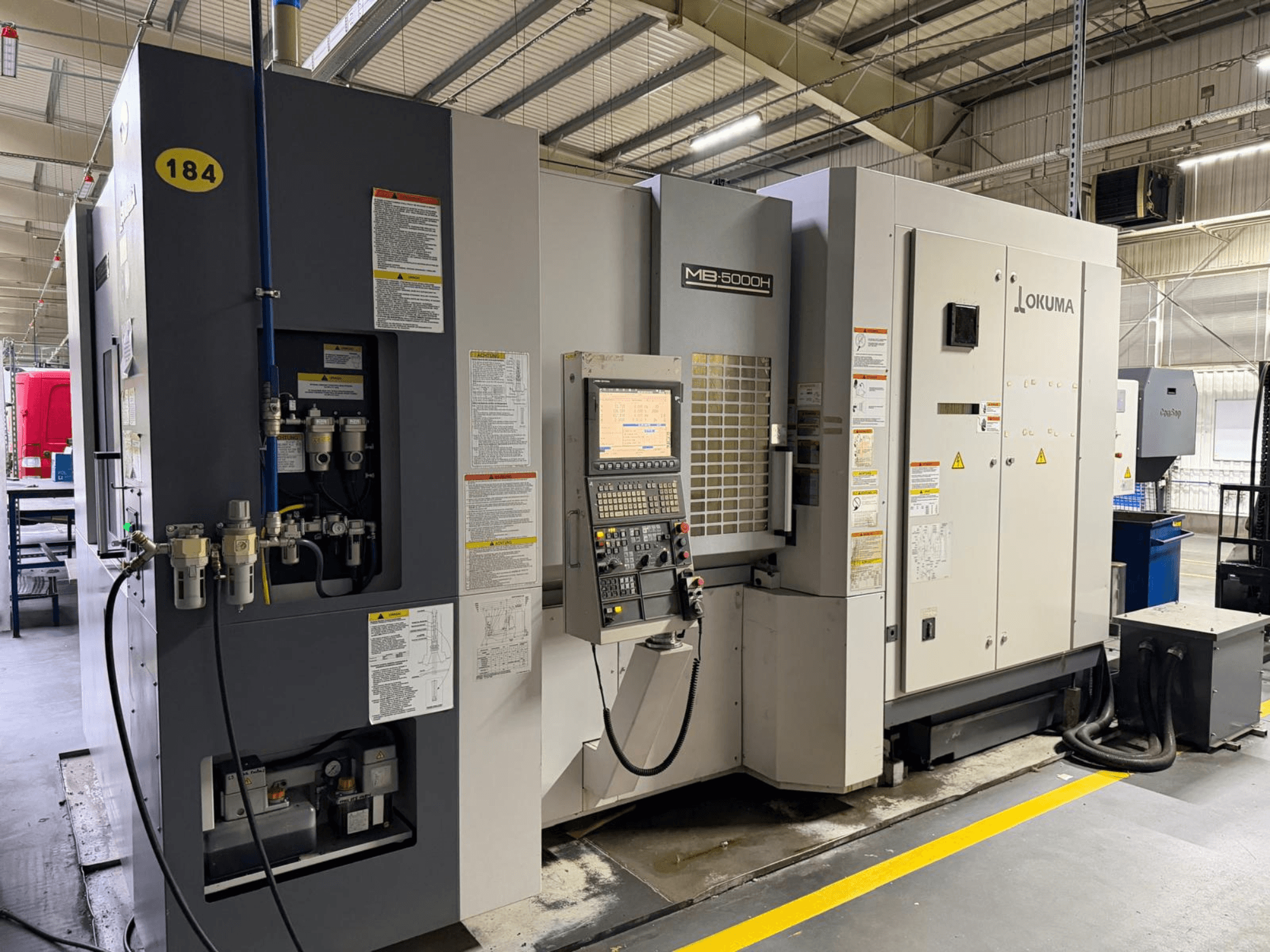 Okuma MB 5000H vízszintes megmunkálóközpont előlnézetben, a kezelőszervek, a feliratozás és a hidraulikus csatlakozások.