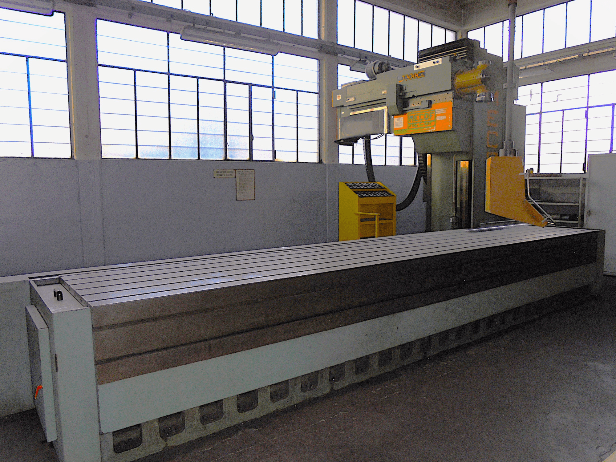 Márkafüggetlen, függőleges marógép egy gyárban, oldalnézetben, nagy, hornyolt felületű asztallal és vezérlőpanellel.