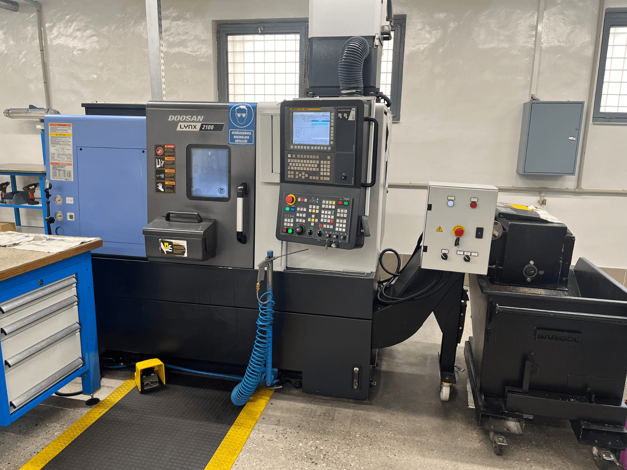 Doosan Lynx 210 eszterga a műhelyben, a vezérlőpanel, a szerszámtároló és a hűtőfolyadék-rendszer, oldalnézet a munkaterület részleteivel.