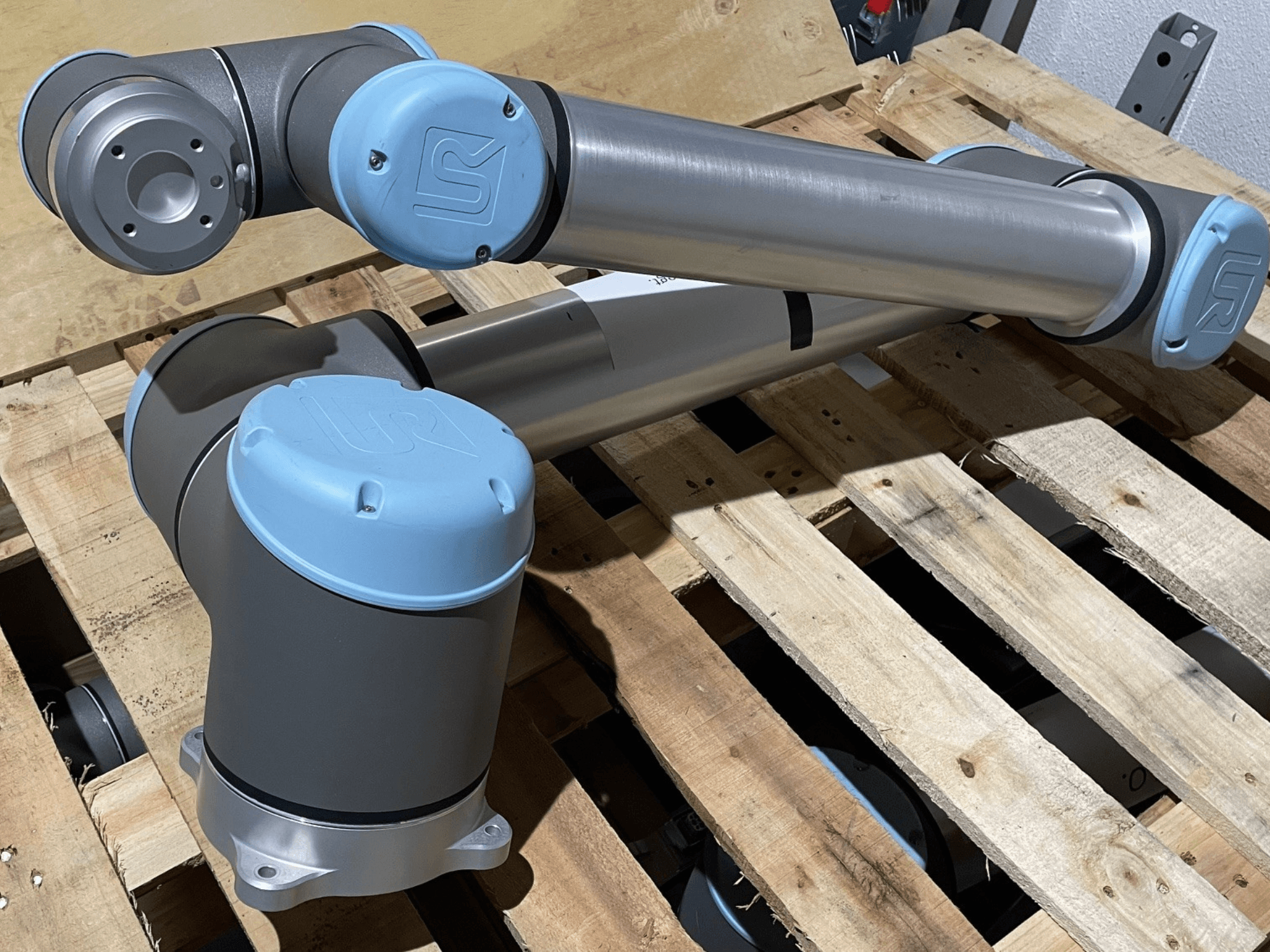 Ezüstszínű robotkar kék végződéssel, elölnézetben, az ízületek és a mechanikai részletek bemutatásával, fából készült raklapon.