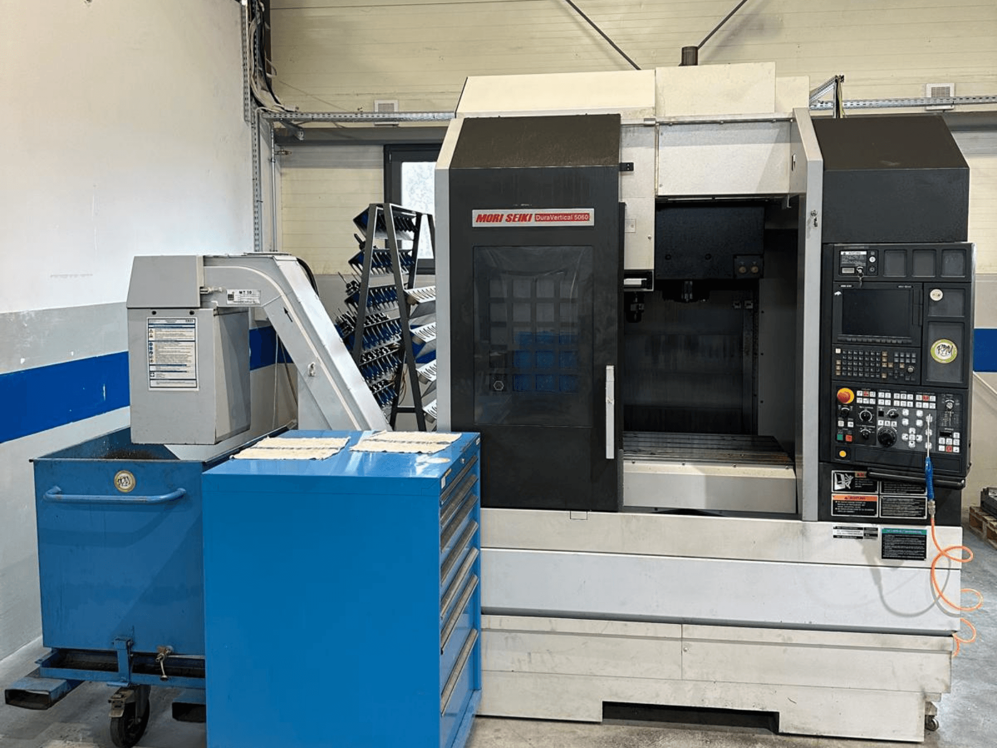 MORI SEIKI DURA VERTICAL 5060 – egy függőleges megmunkáló központ elölnézete a vezérlőpanellel és a közelben elhelyezett szerszámtárolóval.