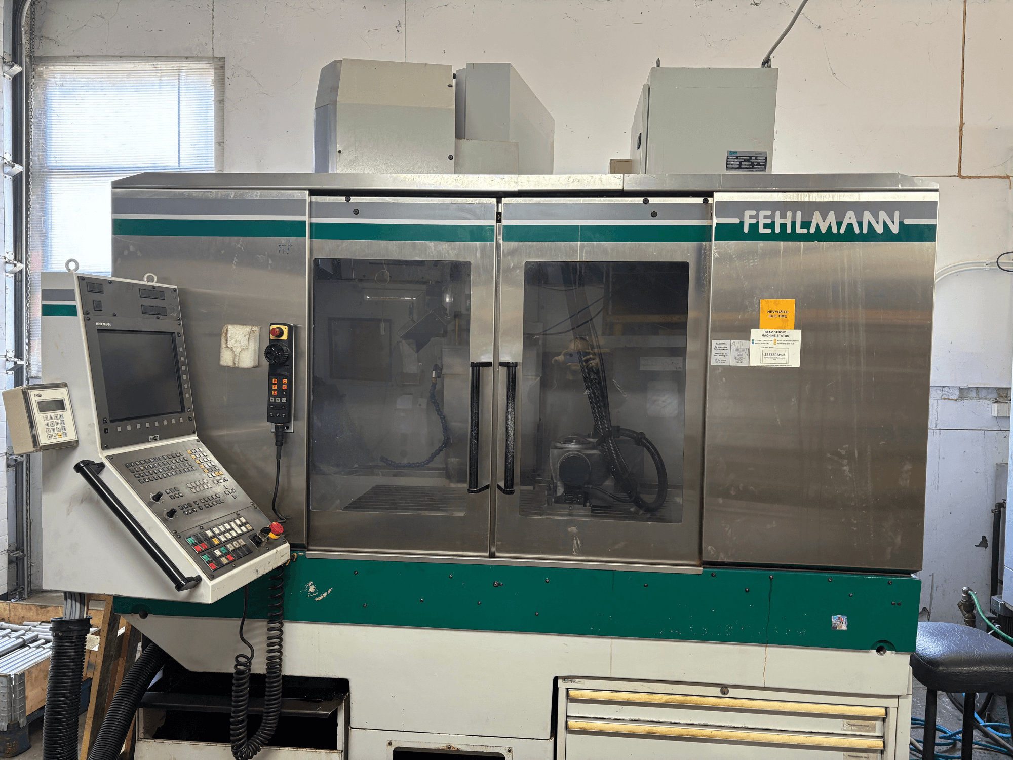A FEHLMANN Picomax 90-M függőleges megmunkáló központ elölnézete, amelyen látható a vezérlőpanel, az átlátszó burkolat és a kezelőgombok.