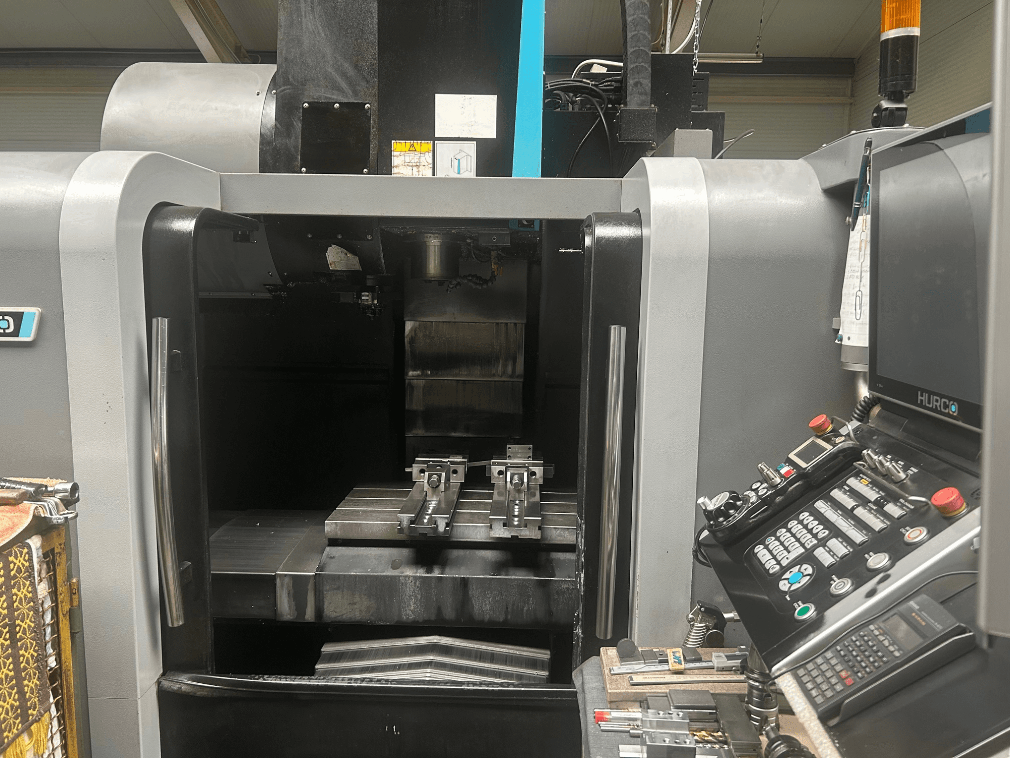 Hurco CNC megmunkálóközpont előlnézetben, szerszámtartó, fém munkadarab, valamint a vezérlőpanel a gombokkal és a kijelzővel.