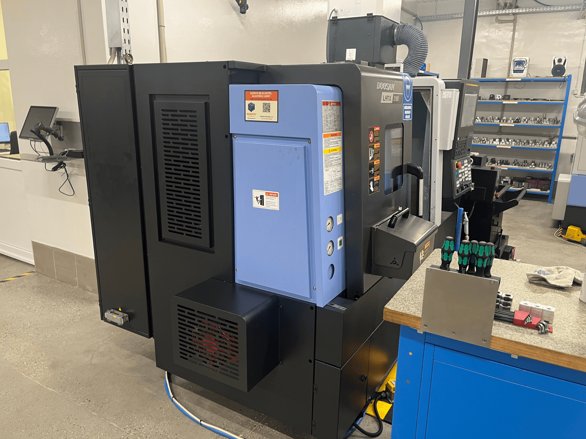 Doosan Lynx 2100 eszterga, oldalnézet, a vezérlőpanel, a szellőzőrendszer és a szerszámok bemutatása a munkapadon.