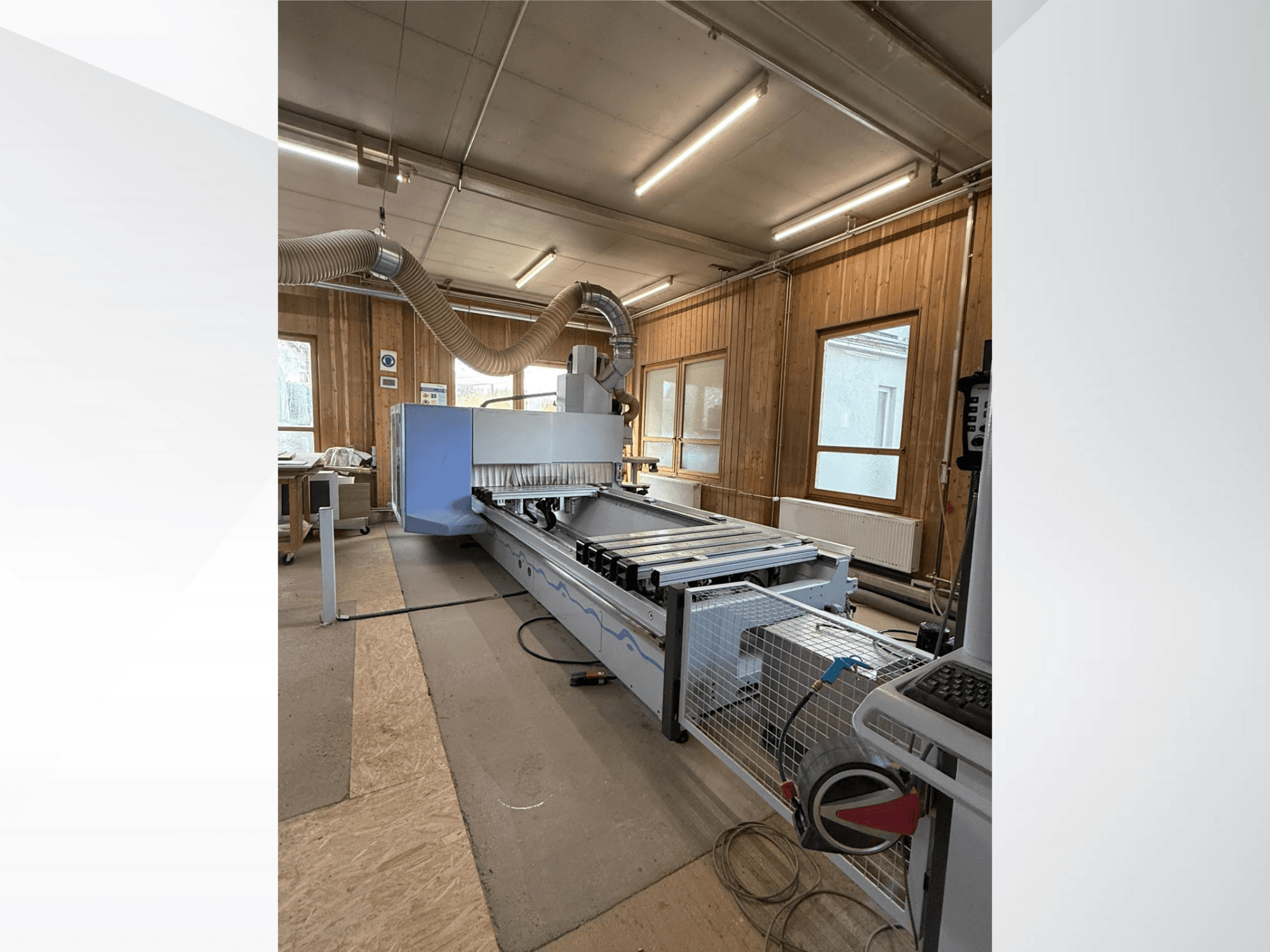 CNC marógép egy műhelyben, oldalnézetben, nagy kék testtel, csatlakoztatott szalagszállítóval és látható vezérlőpanellel.