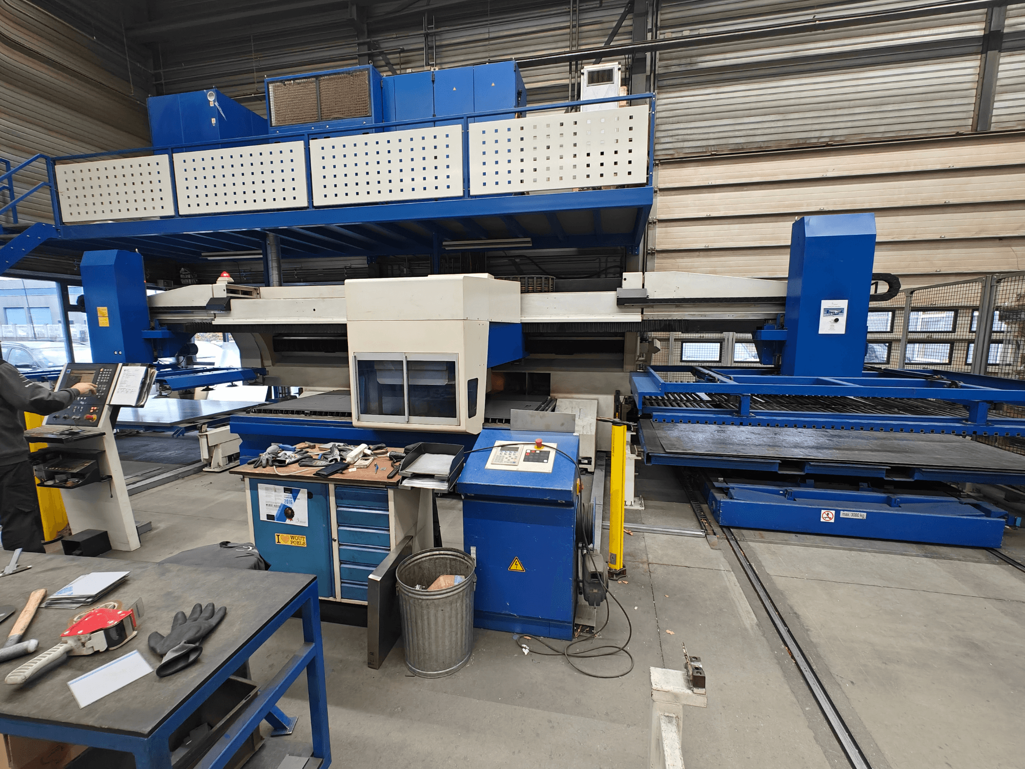 Ipari CNC-gép elölnézetből, kék-fehér szerkezettel, vezérlőpanellel és különböző szerszámokkal a munkaasztalon.