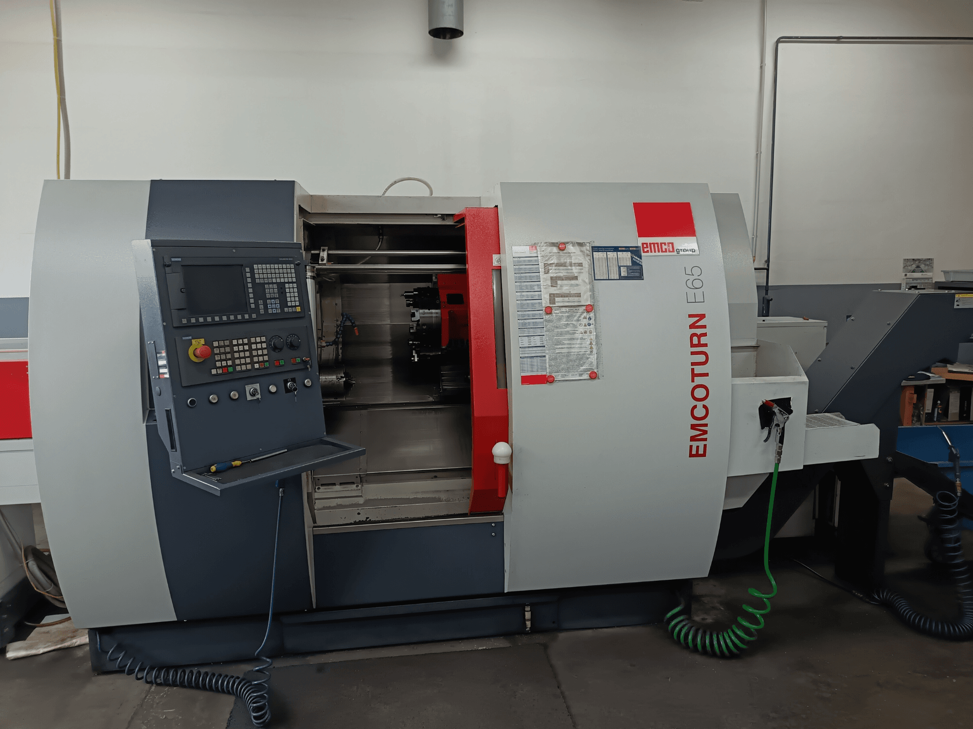 EMCO EMCOTURN E65 esztergapadozó központ, előlnézet, amely a vezérlőpanelt, a belső mechanizmust és a műhelyben lévő funkciókat mutatja.