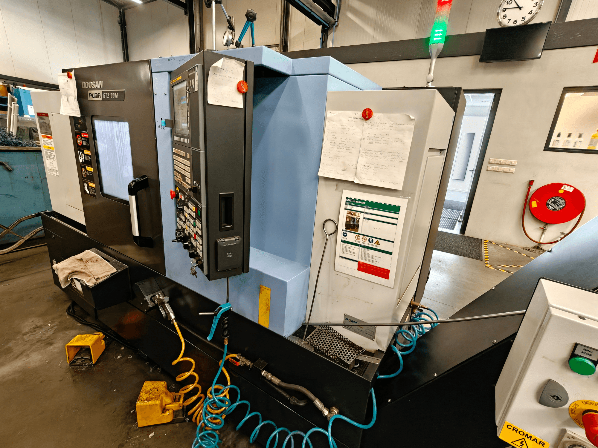 A DOOSAN Puma GT 2100M vízszintes esztergagép elülső nézete a vezérlőpanellel, a biztonsági berendezésekkel és a karbantartási jegyzetekkel.