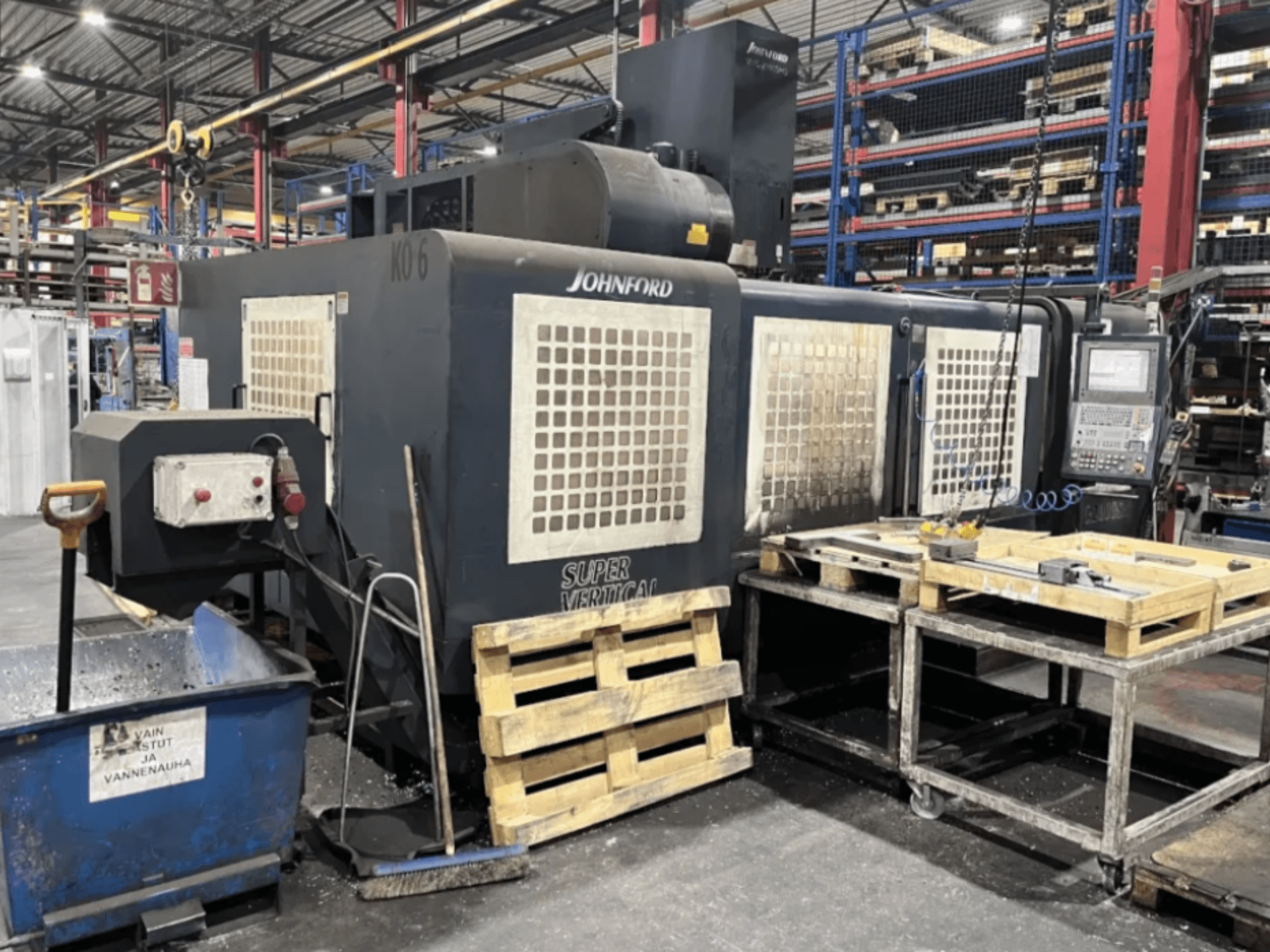 Johnford Super Vertical marógép egy gyárban, oldalnézet a vezérlőpanellel, raklapokkal és tisztítóberendezéssel a közelben.