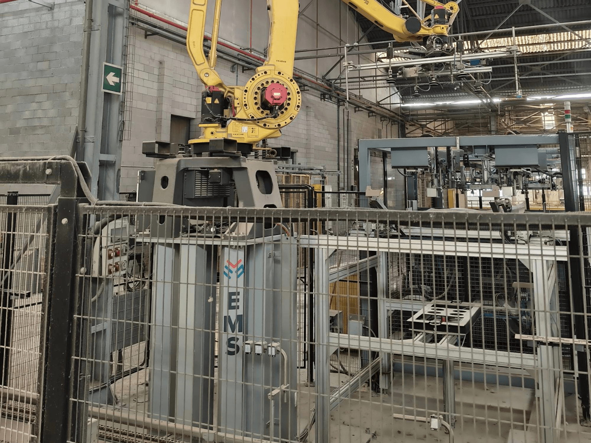 A FANUC M-410iC/315 robotkar elölnézete, amely egy EMS-szerszám felett helyezkedik el, a háttérben ipari berendezésekkel és védőkerítéssel.