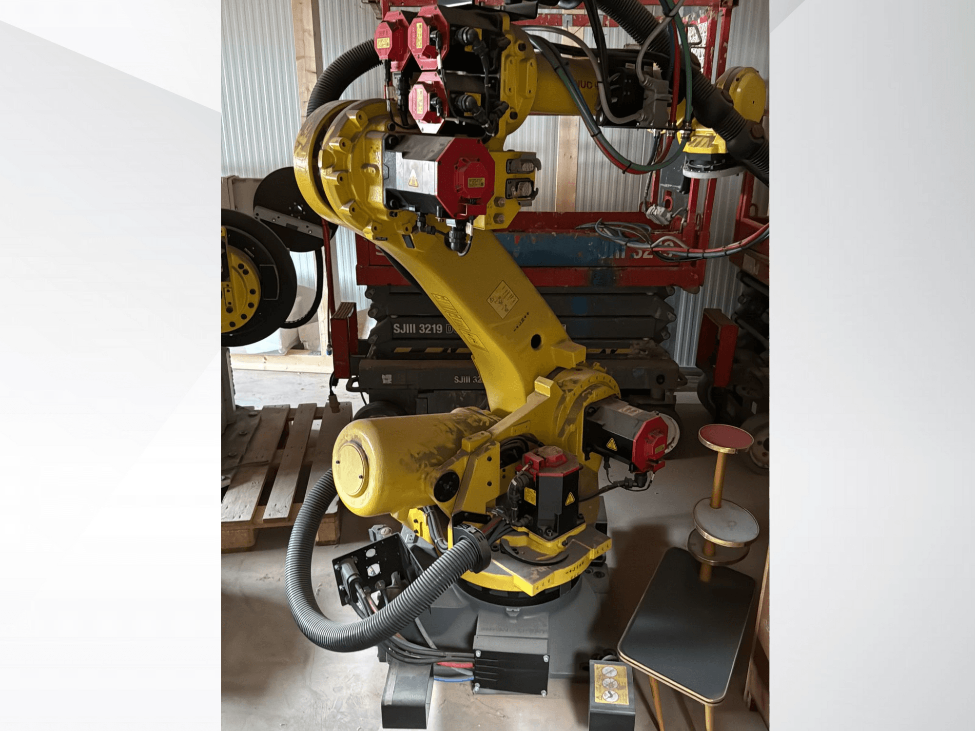 Sárga ipari robotkar piros vezérlőkkel, elölnézetben; több ízülettel és tömlőkkel rendelkezik, egy munkaterületen állítva.