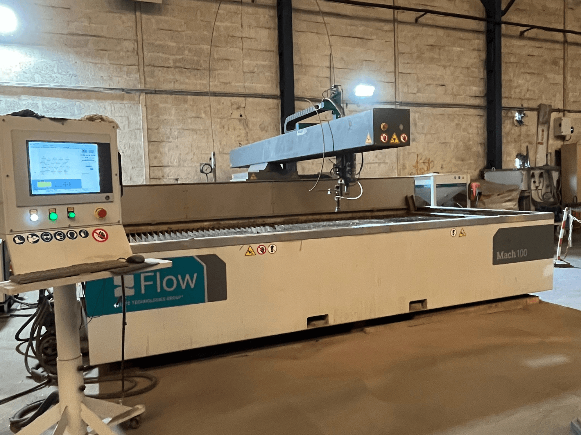 Flow Mach 100 vízsugaras vágógép, oldalnézet, digitális vezérlőpanel és vágófej látható a munkaasztalon.