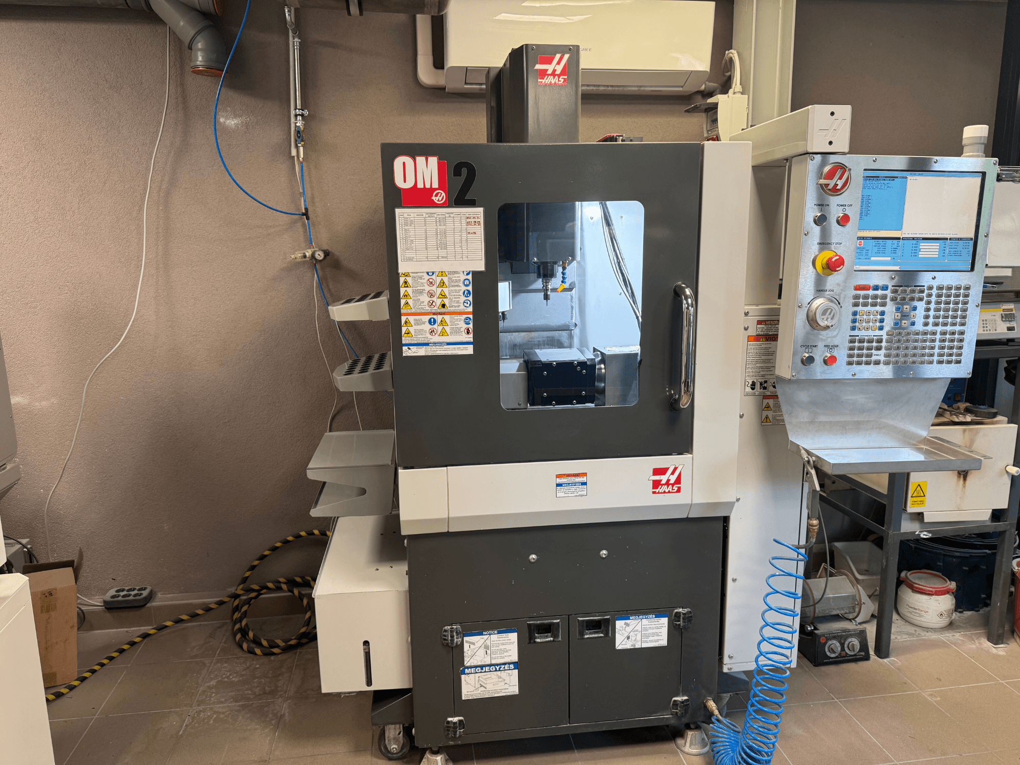 Haas OM-2 CNC-gép, elölnézet, vezérlőpanel, orsó és biztonsági címkék, műhelykörnyezetben.