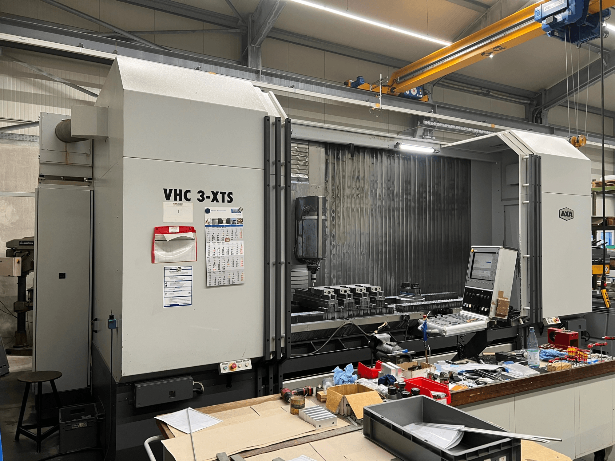 AXA VHC 3-XTS CNC-gép oldalnézetben, vezérlőpanel és szerszámokkal és alkatrészekkel zsúfolt munkaterület.