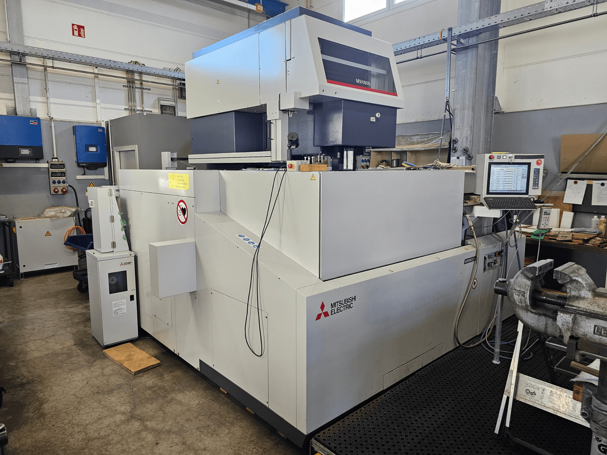 Mitsubishi MV4800 huzalos szikraforgácsoló gép egy műhelyben, oldalnézetben a vezérlőpanel, a biztonsági címkék és a környező berendezések.