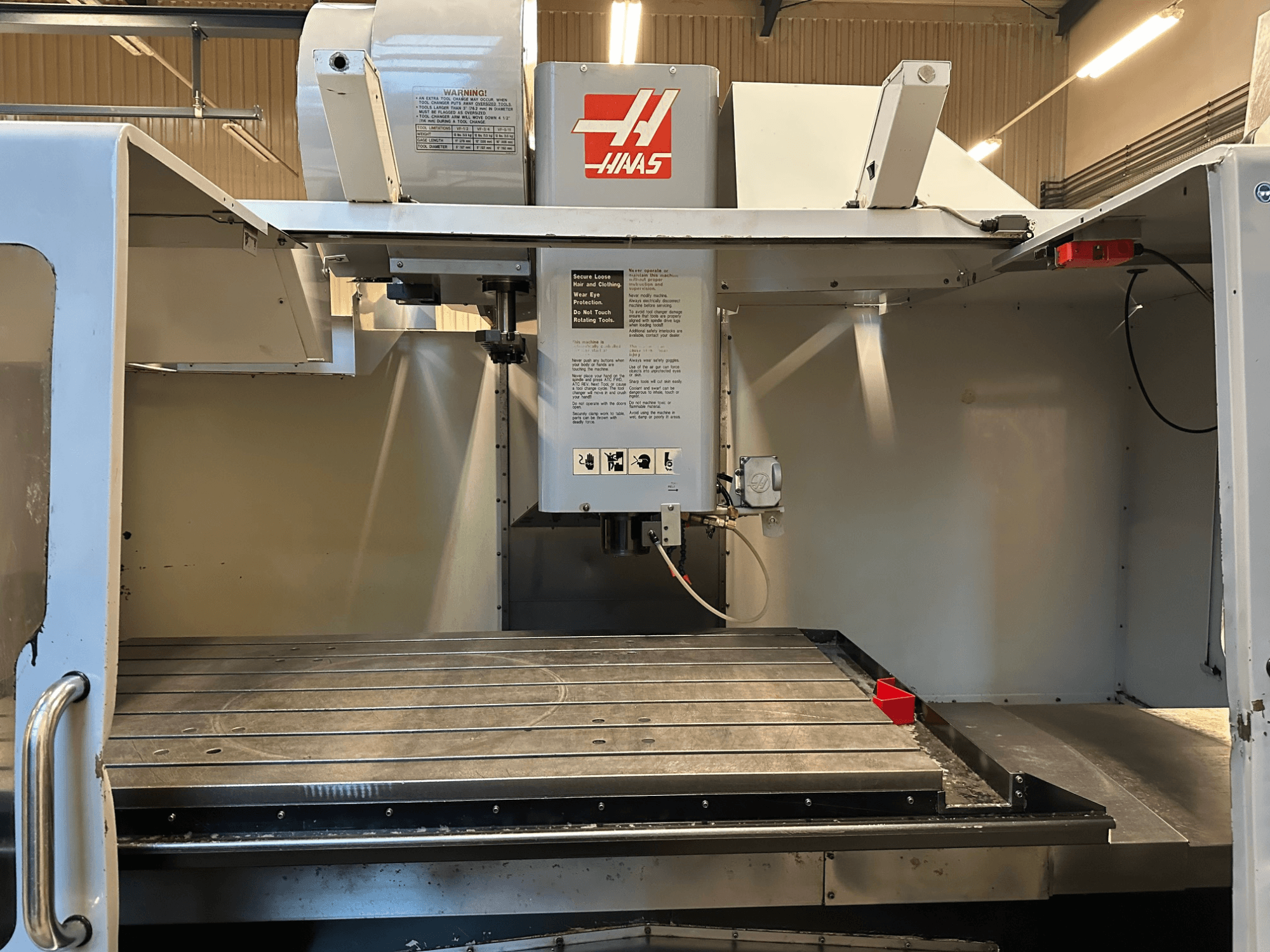 Haas CNC marógép, VF-2 modell, elölnézetben a kezelőszervek, a fém munkaasztal és a biztonsági címke a panelen.