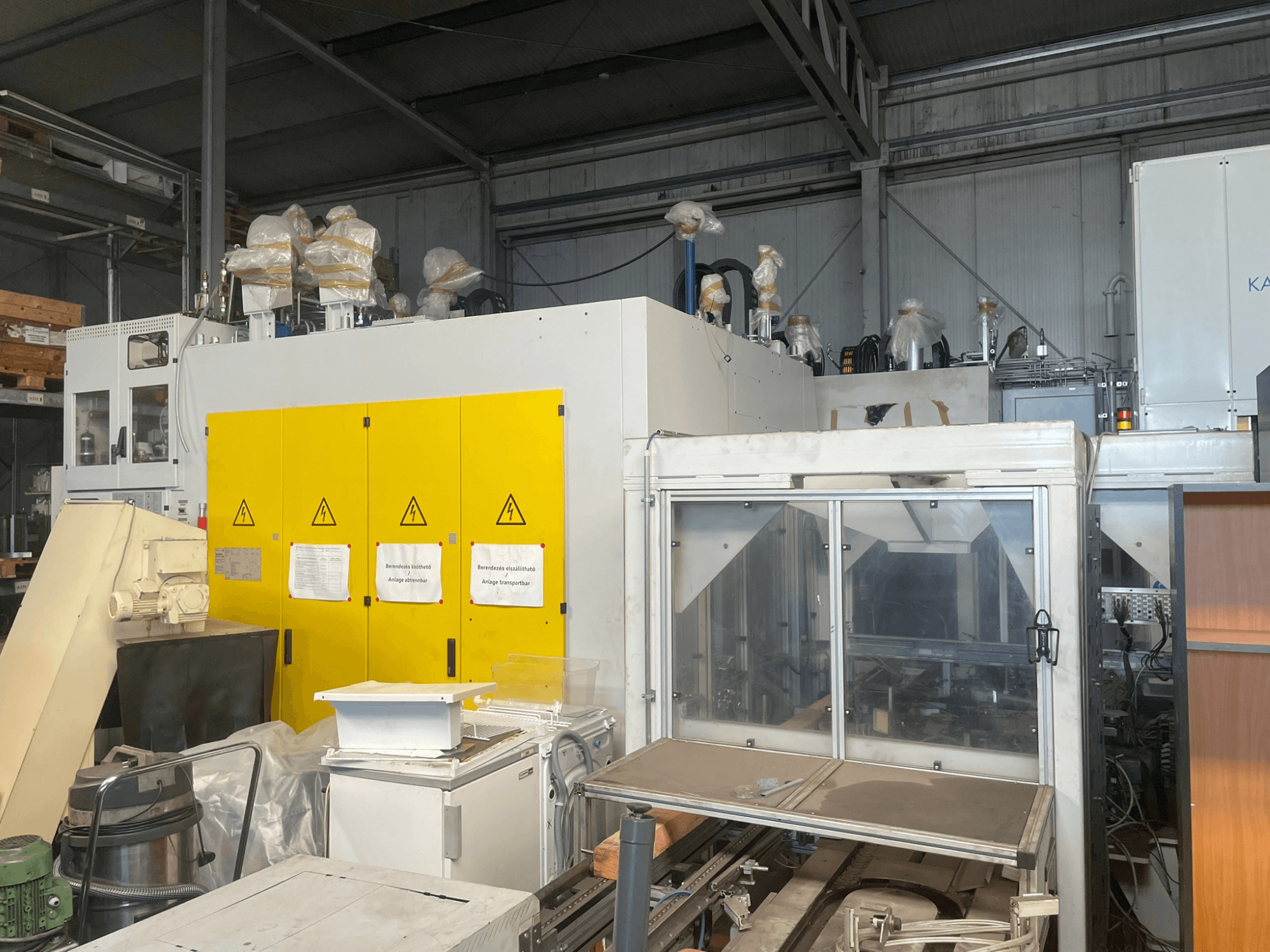 Ipari gépek sárga vezérlőpanellel, oldalnézetben, figyelmeztető címkékkel és műanyag borítással, raktári környezetben.