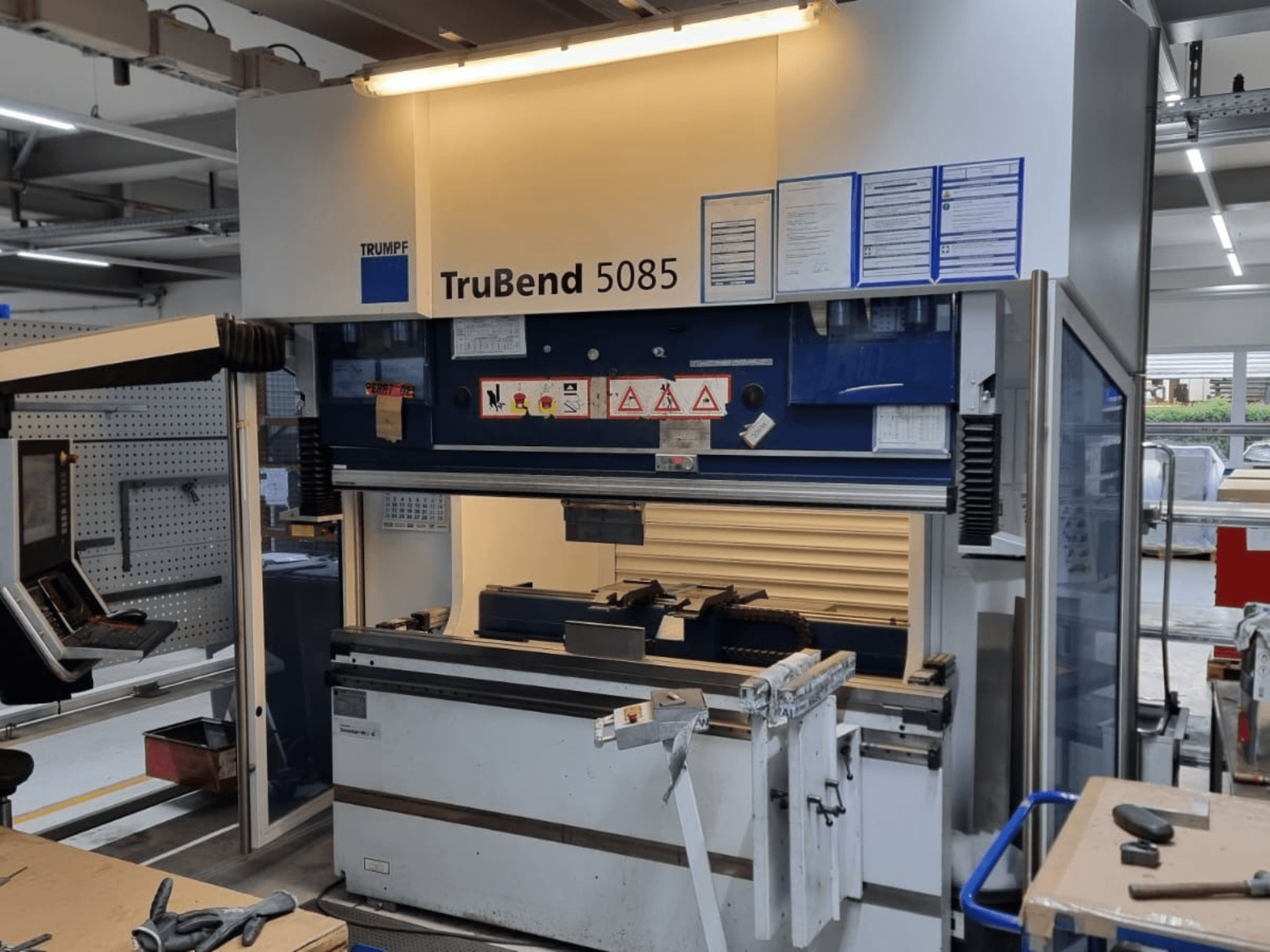 TRUMPF TruBend 5085 hajlítógép, előlnézet, a műhelyben lévő kezelőpanel, kezelőállomás és biztonsági figyelmeztetések.
