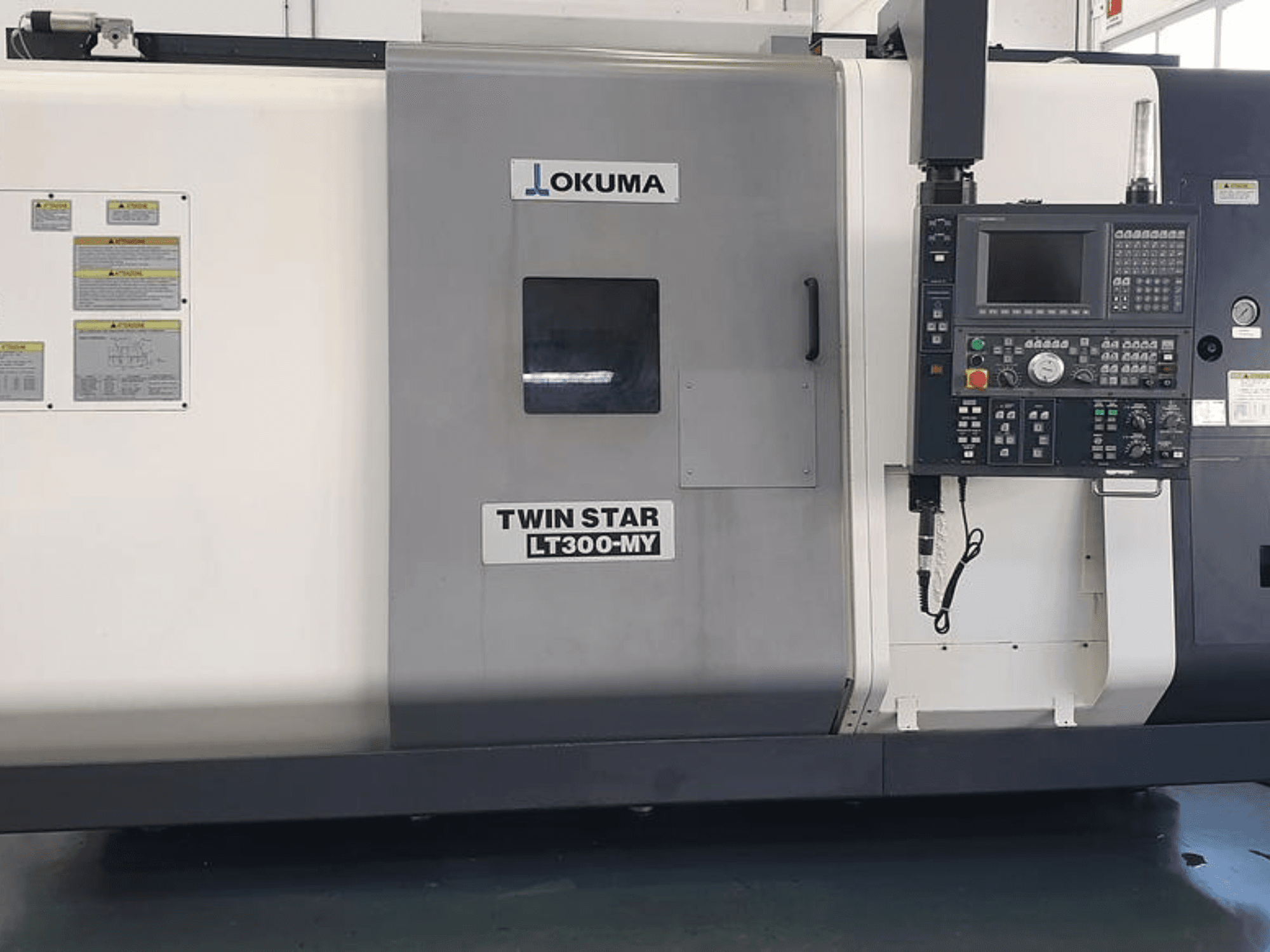 Okuma Twin Star LT300-MY CNC eszterga oldalnézetben, a vezérlőpanel, a kezelőgombok és a biztonsági címkék.