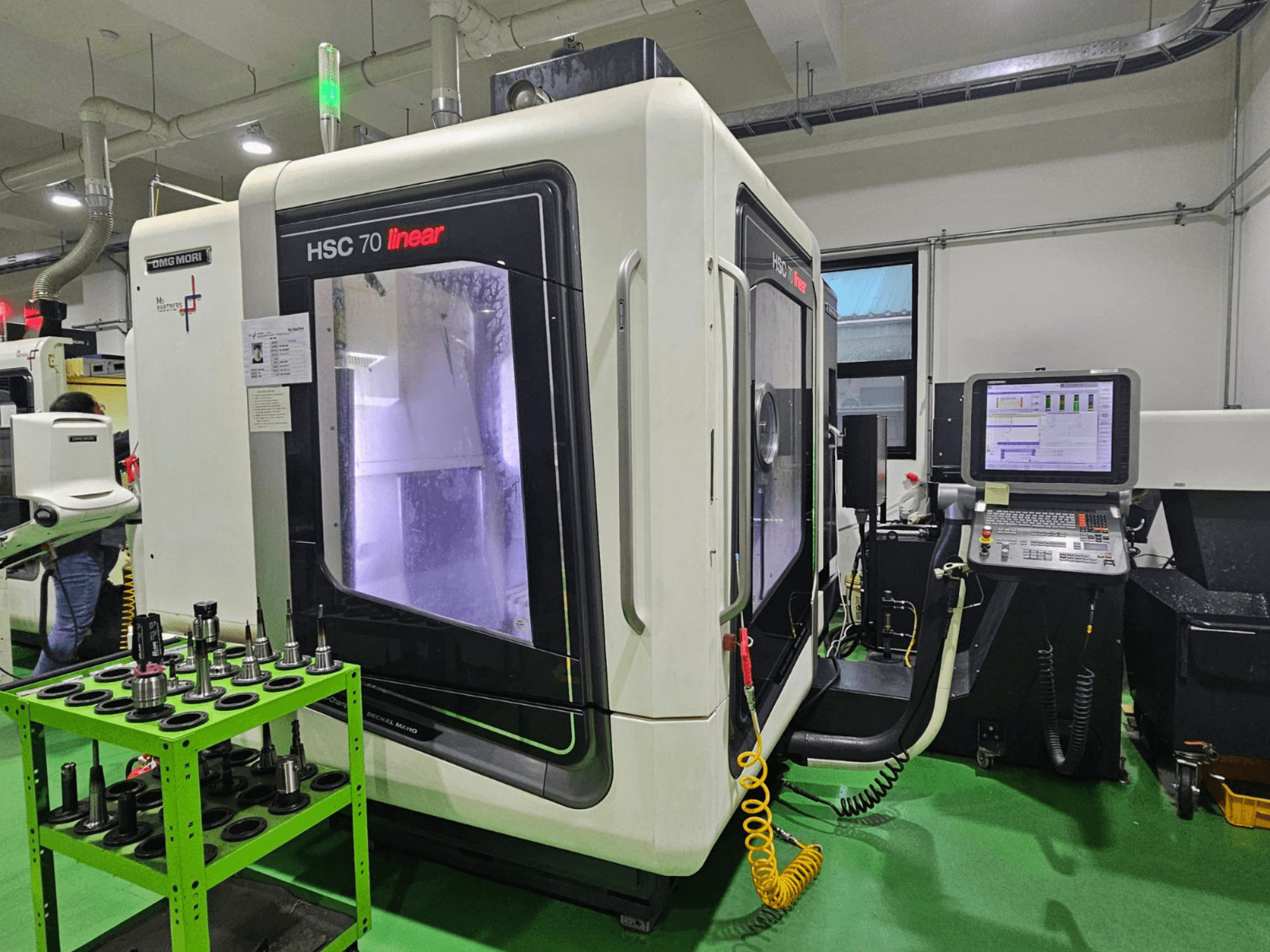 DMG MORI HSC 70 lineáris univerzális megmunkálóközpont, elölnézet, szerszámtartókkal a zöld asztalon és látható a vezérlőpanel.