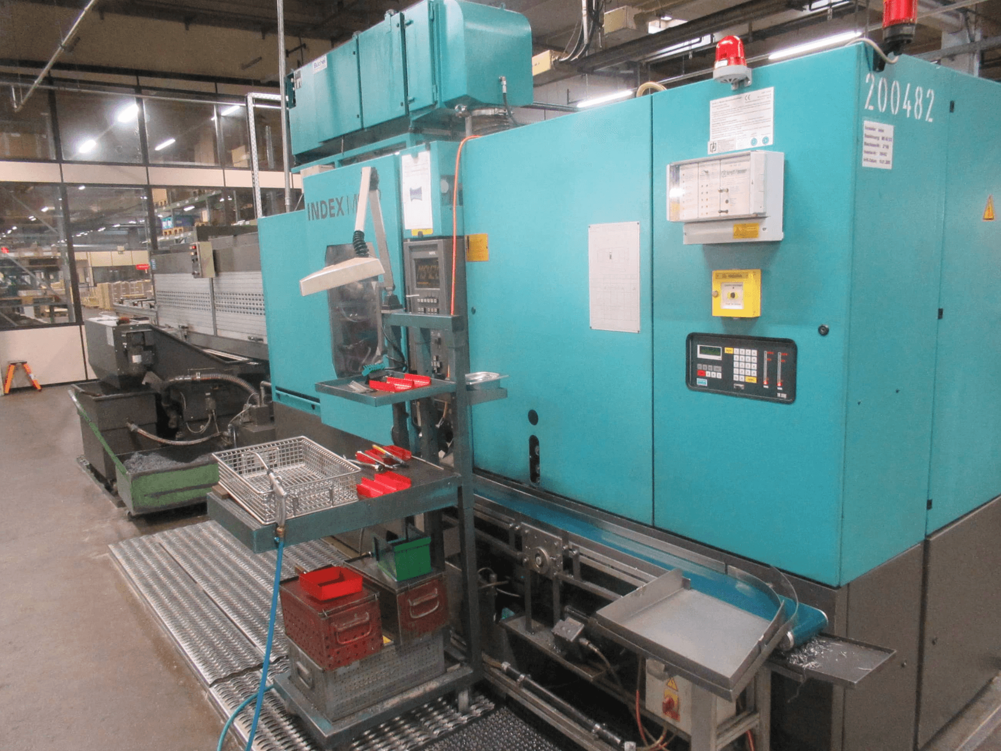 Türkiz INDEX gép a gyárban, oldalnézet; vezérlőpanel, szerszámtároló és szállítószalag. Látható tárolók, tömlők és riasztók.