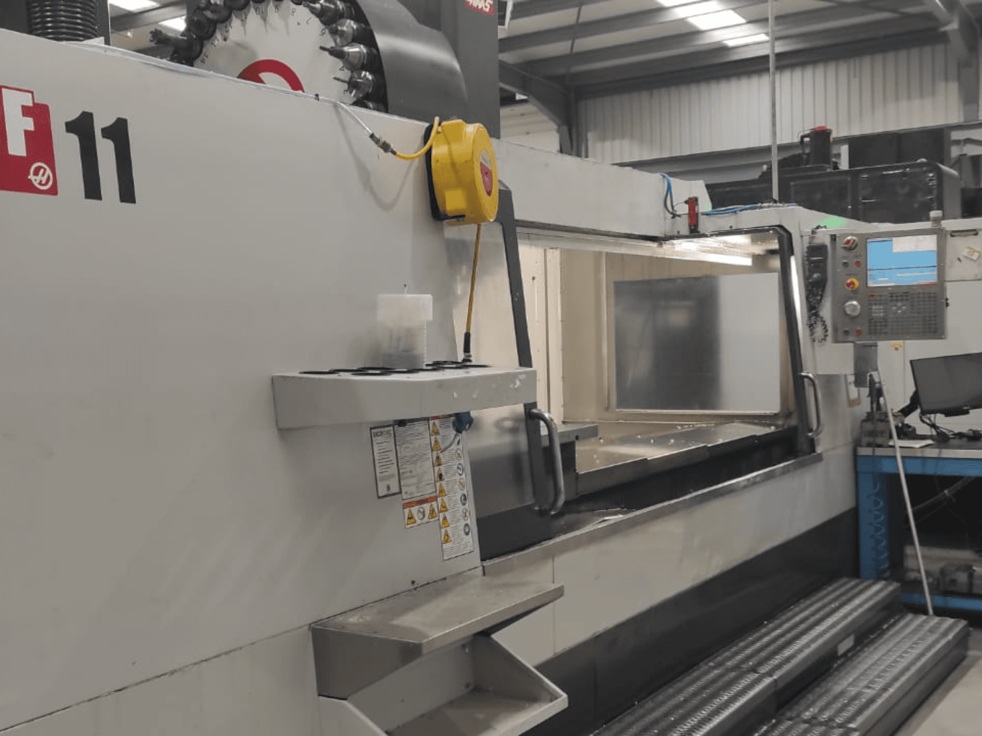 A Haas VF-11 típusú gépe elölnézetből, a vezérlőpanel, a nagy munkaterület és a biztonsági címkék láthatóak.