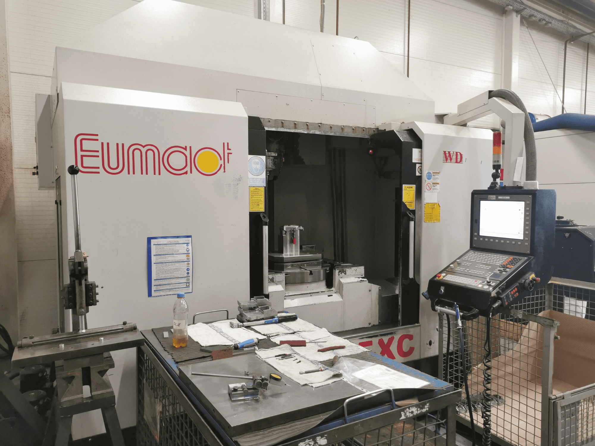 Egy Eumdaot CNC-gép előlapja, vezérlőpanellel, szerszámokkal, valamint alkatrészekkel és anyagokkal zsúfolt munkafelülettel.