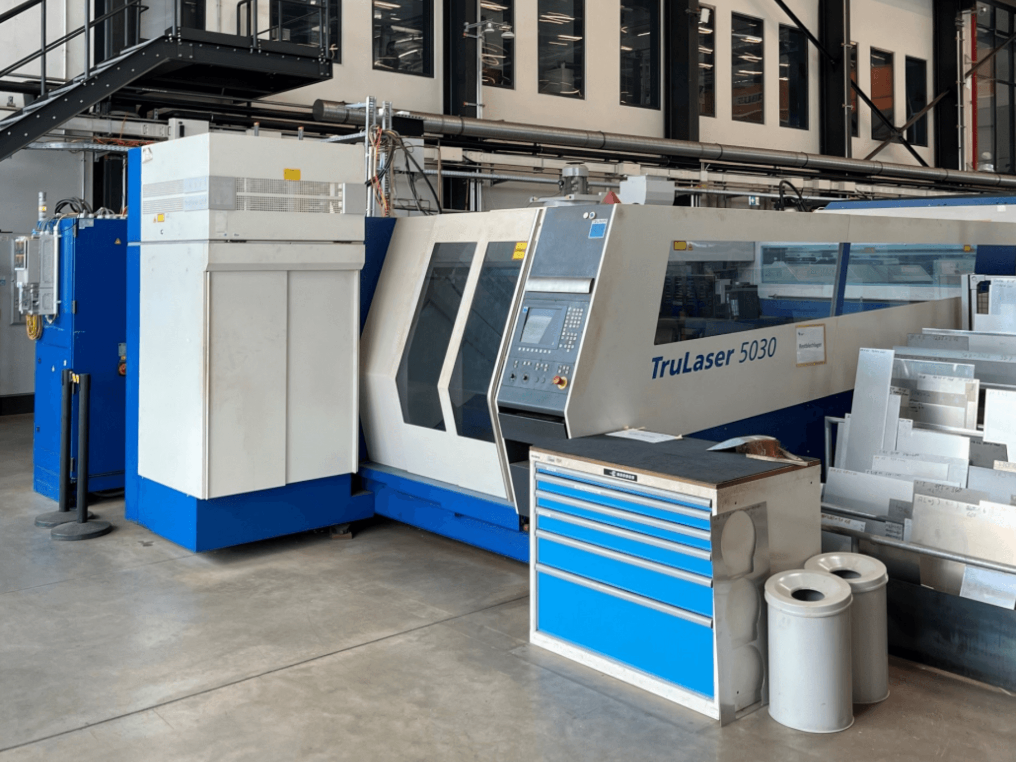 A TRUMPF TruLaser 5030 CO₂ lézervágógép elölnézetből, a vezérlőpanellel és a mellette lévő, fémlemezekkel ellátott munkapaddal.