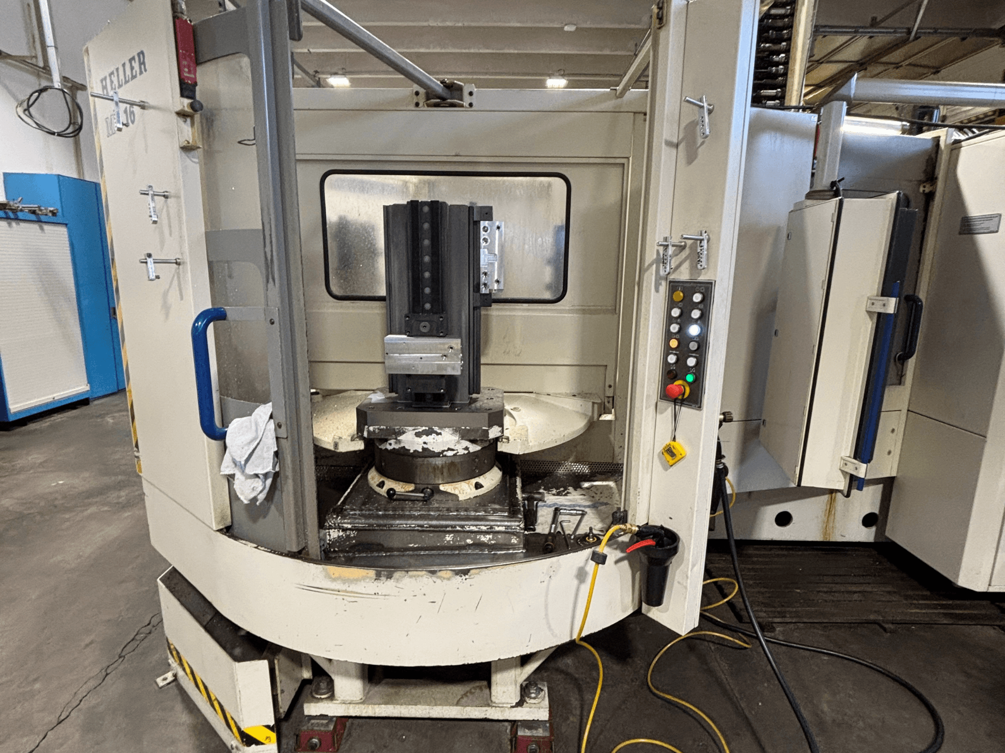 Heller megmunkálóközpont, függőleges modell, elölnézetben az orsó, a vezérlőpanel és a munkaasztal a rögzített berendezésekkel.