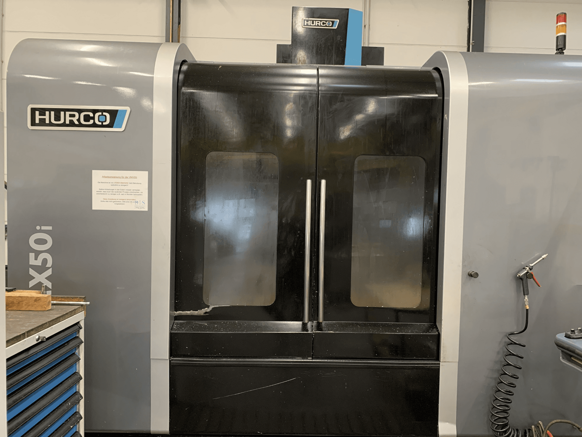 Egy Hurco VX50i CNC-gép előlnézetből, kettős ajtóval, műszaki kezelőszervekkel és egy karbantartási táblával az oldalán.