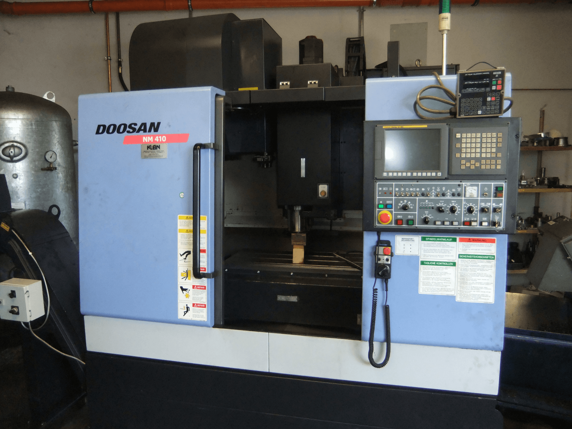 DOOSAN NM 410 függőleges megmunkáló központ, elölnézet, amelyen látható a vezérlőpanel, az orsóterület és a biztonsági címkék.