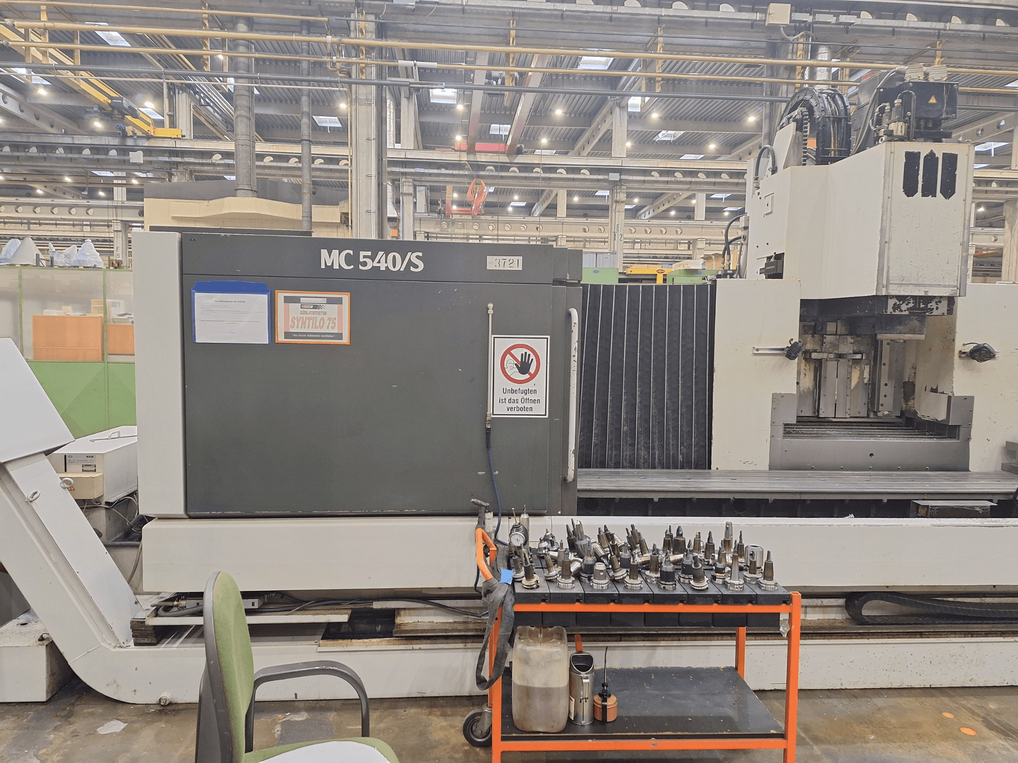 Függőleges megmunkálóközpont STAMA MC 540S egy műhelyben, oldalnézet, szerszámtartókkal egy kocsin a közelben.