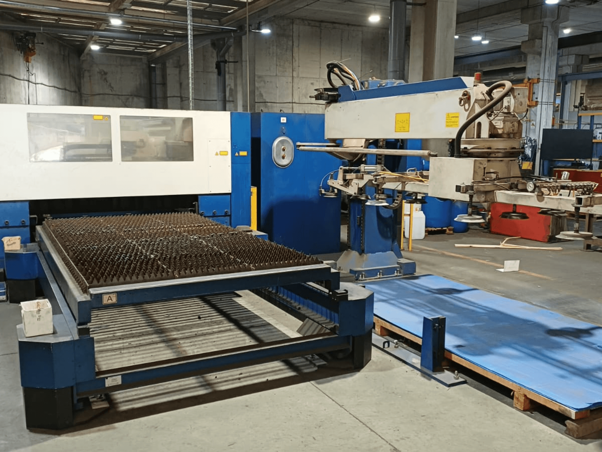 TRUMPF TRUMATIC L 3030 CO₂-lézervágógép, oldalnézet, amelyen látható a tüskés felületű vágóasztal és a robotkar.