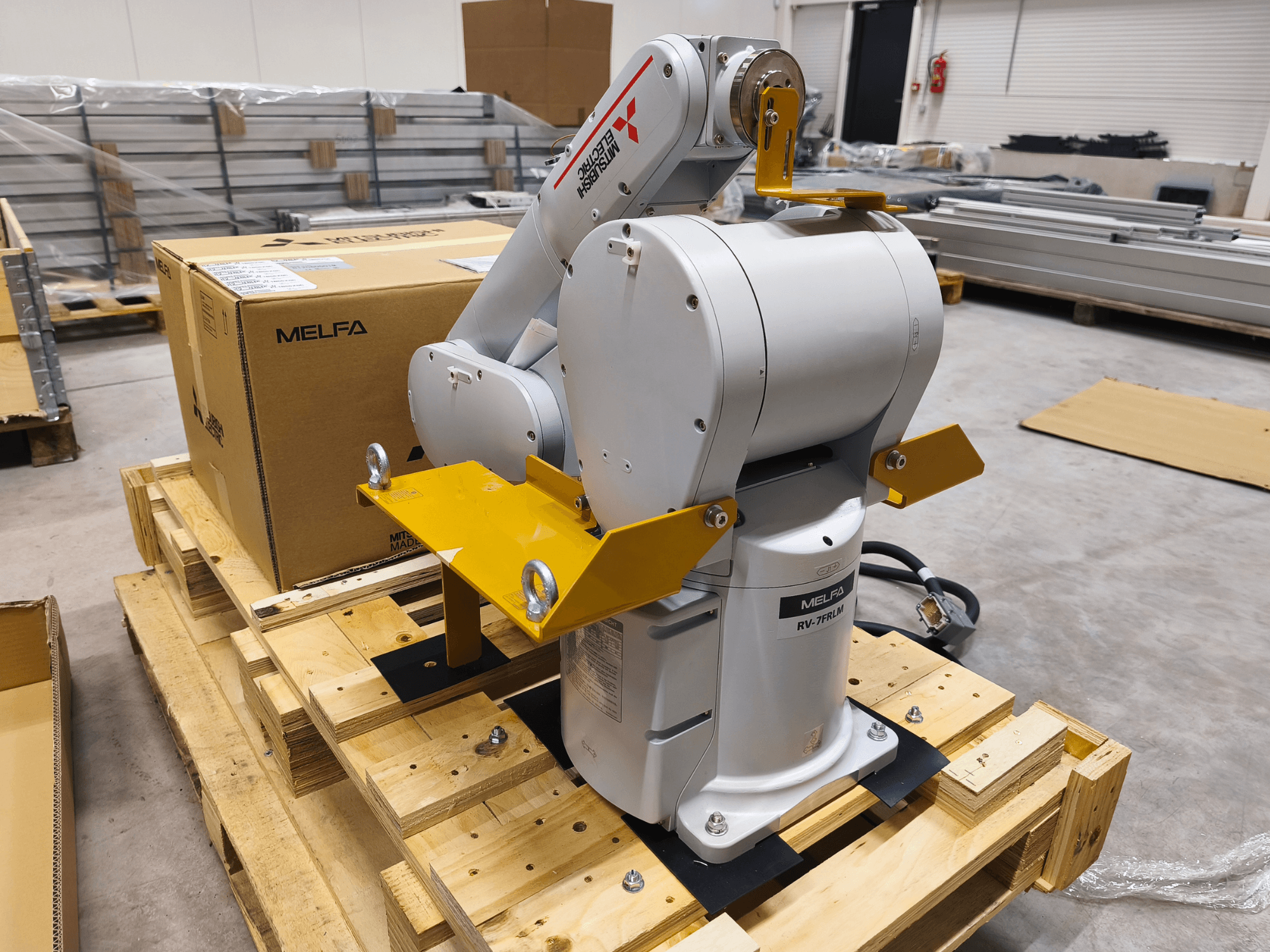 MELFA RV-7FLM ipari robotkar fa raklapon, elölnézetben, sárga tartó konzolokkal és szürke csomagoló dobozban.