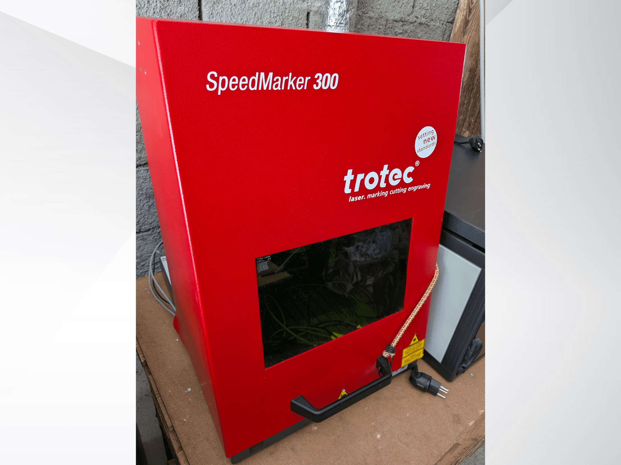 Piros Trotec SpeedMarker 300 lézeres jelölőgép elölnézetben, nagyméretű ablakkal és hálózati csatlakozóval.